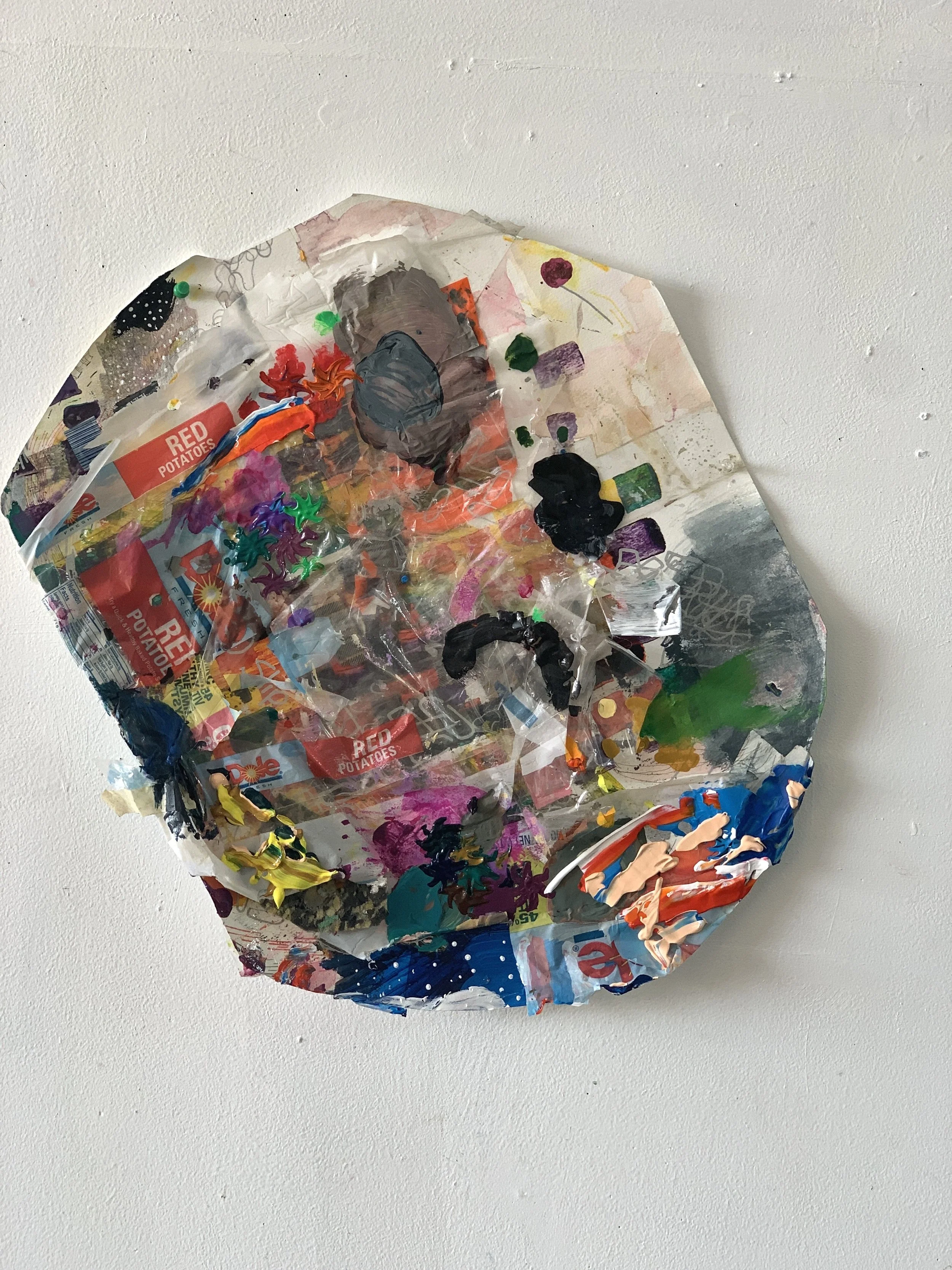 A crumpled and painted piece of aluminum foil with various colors and textures, including black, green, purple, yellow, and red, placed on a white wall.
