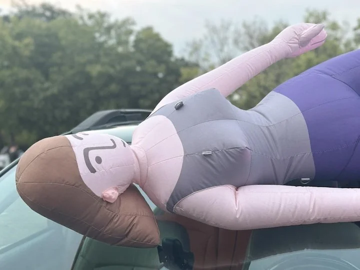 A large inflatable doll with a cartoonish face, long brown hair, wearing a gray top and purple skirt, lying on a car's hood.