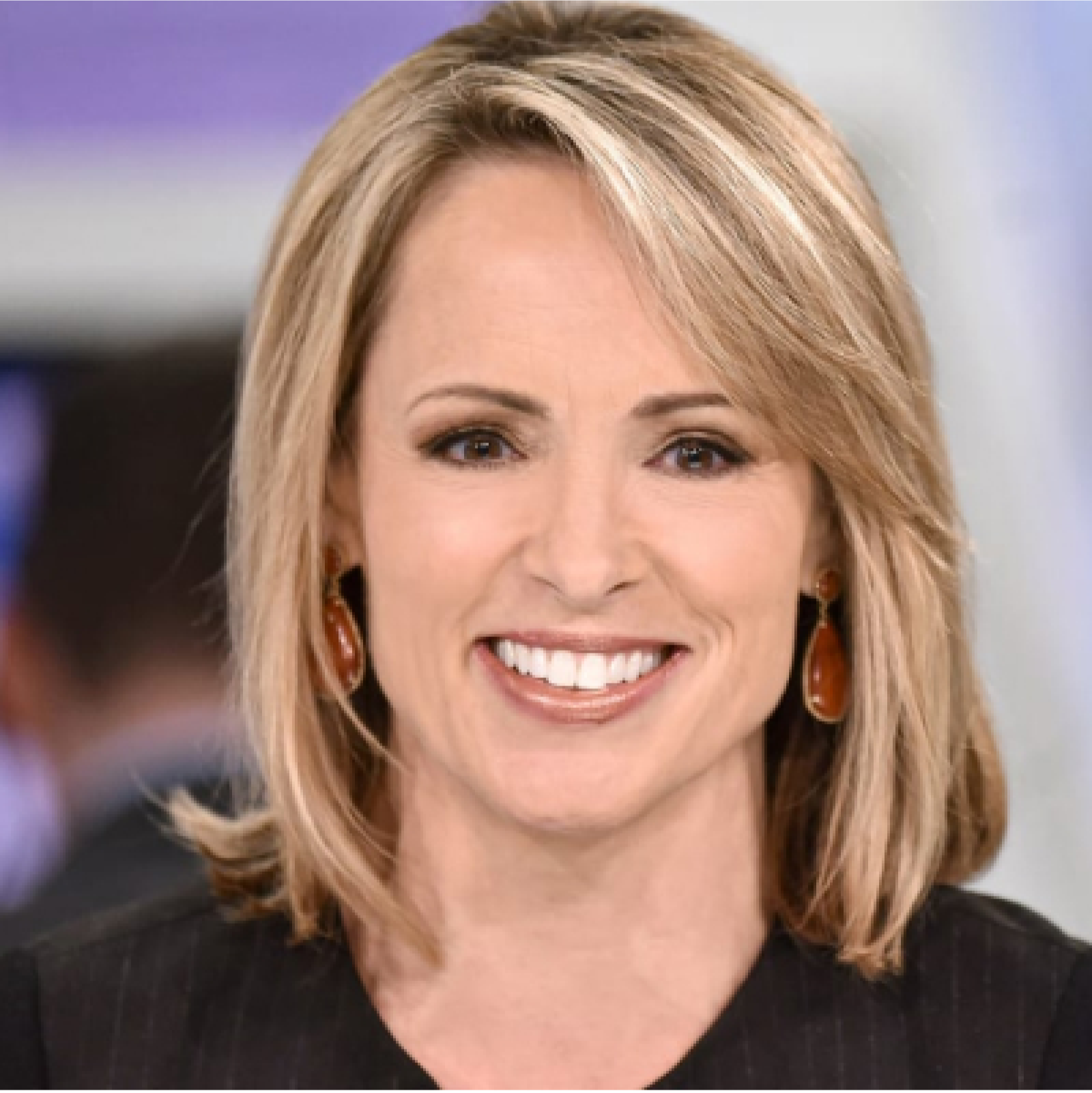 Close-up of a woman with blonde hair smiling, wearing earrings and a dark blazer, in an indoor setting.