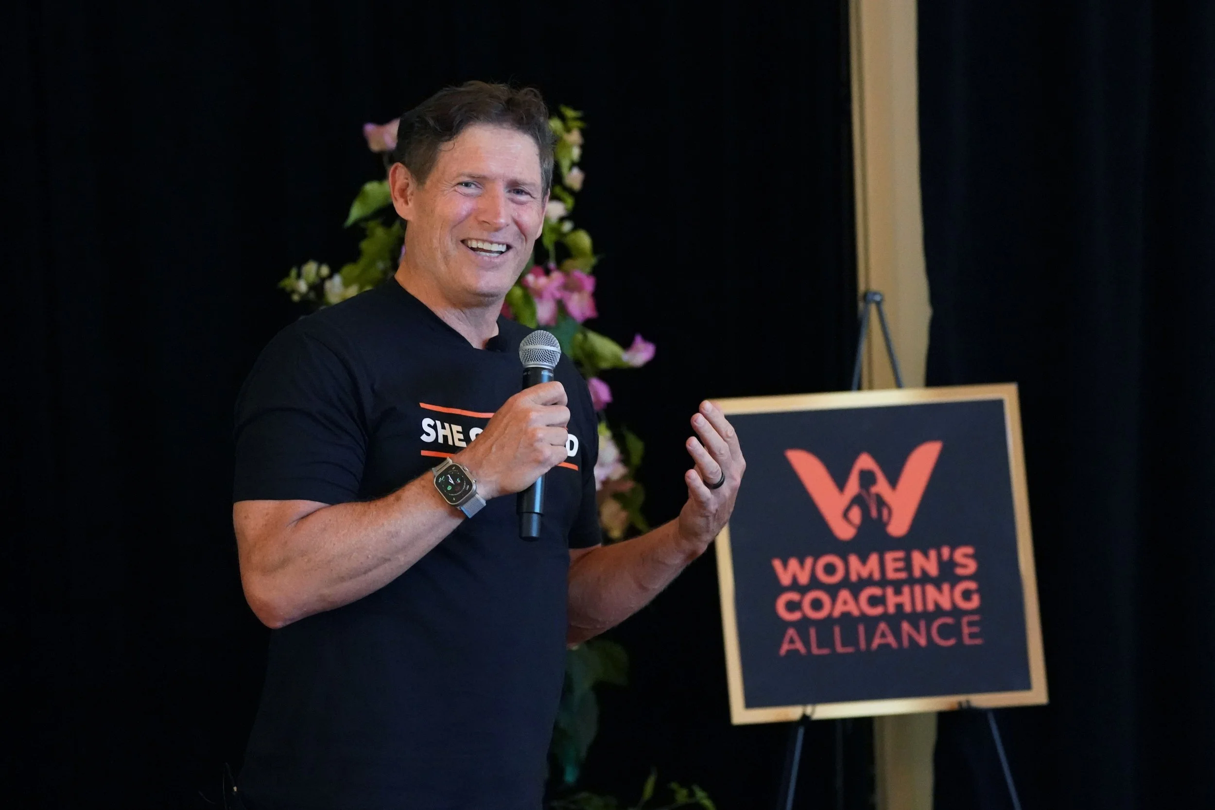 Man speaking at an event with a Women’s Coaching Alliance sign in the background.