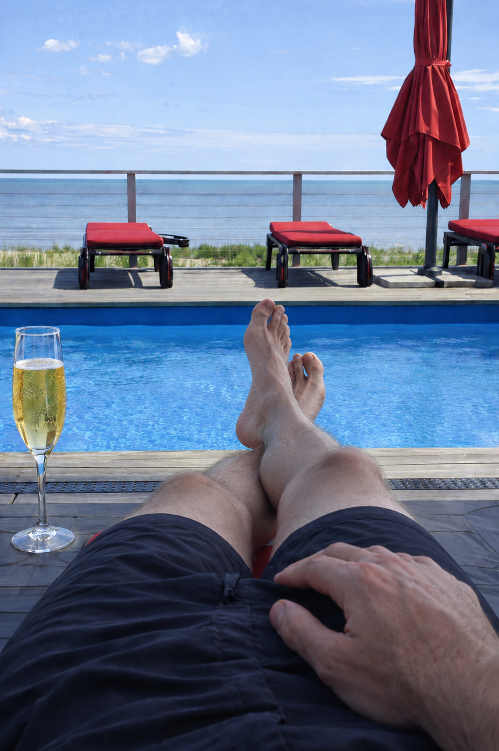 Person relaxing by a pool with legs crossed, holding a hand on their stomach, and a glass of champagne on the poolside. Pool chairs and a red umbrella are visible on the deck, overlooking the ocean under a partly cloudy sky.