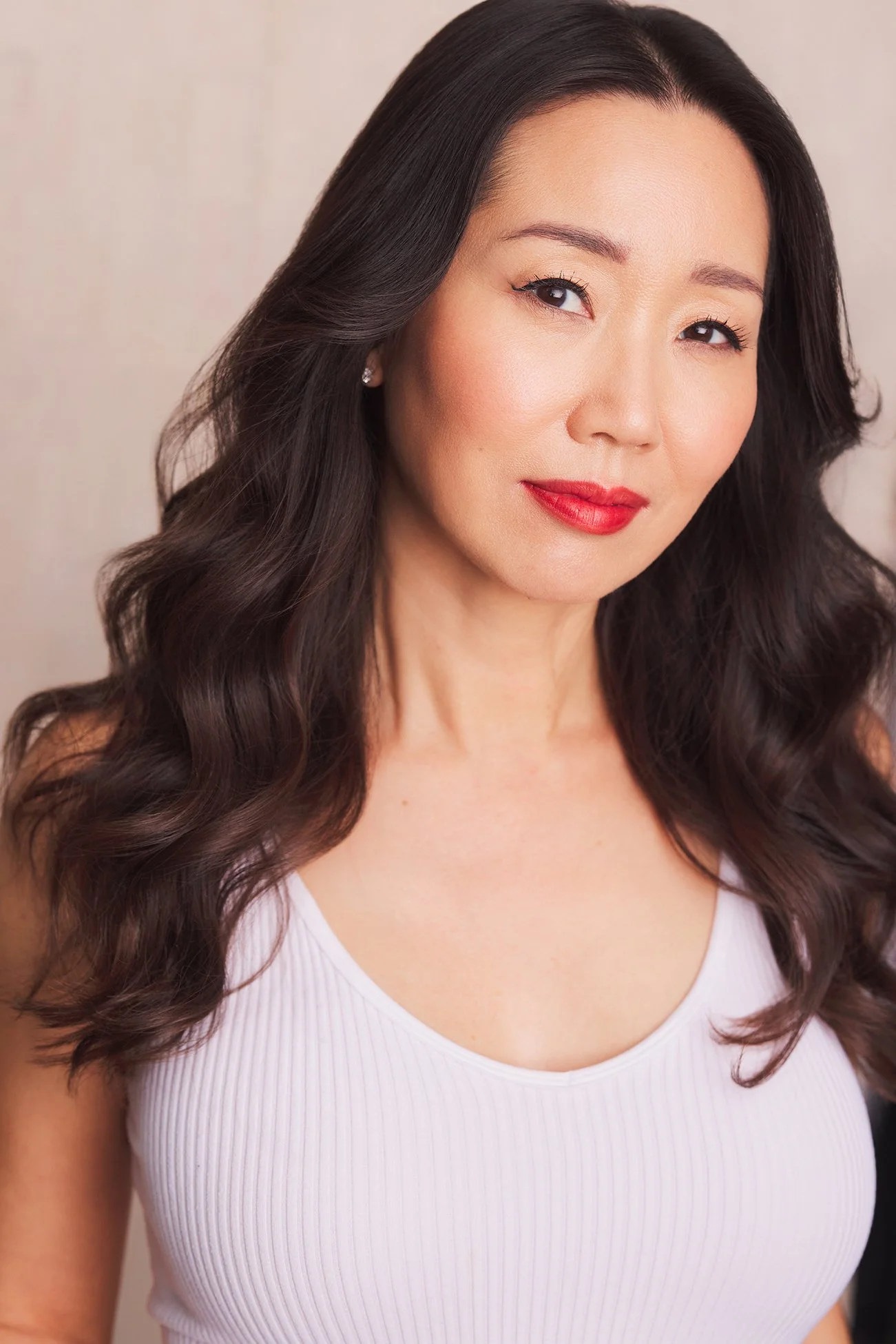 A woman with long dark hair, red lipstick, wearing a white sleeveless top, smiling slightly, against a neutral background.