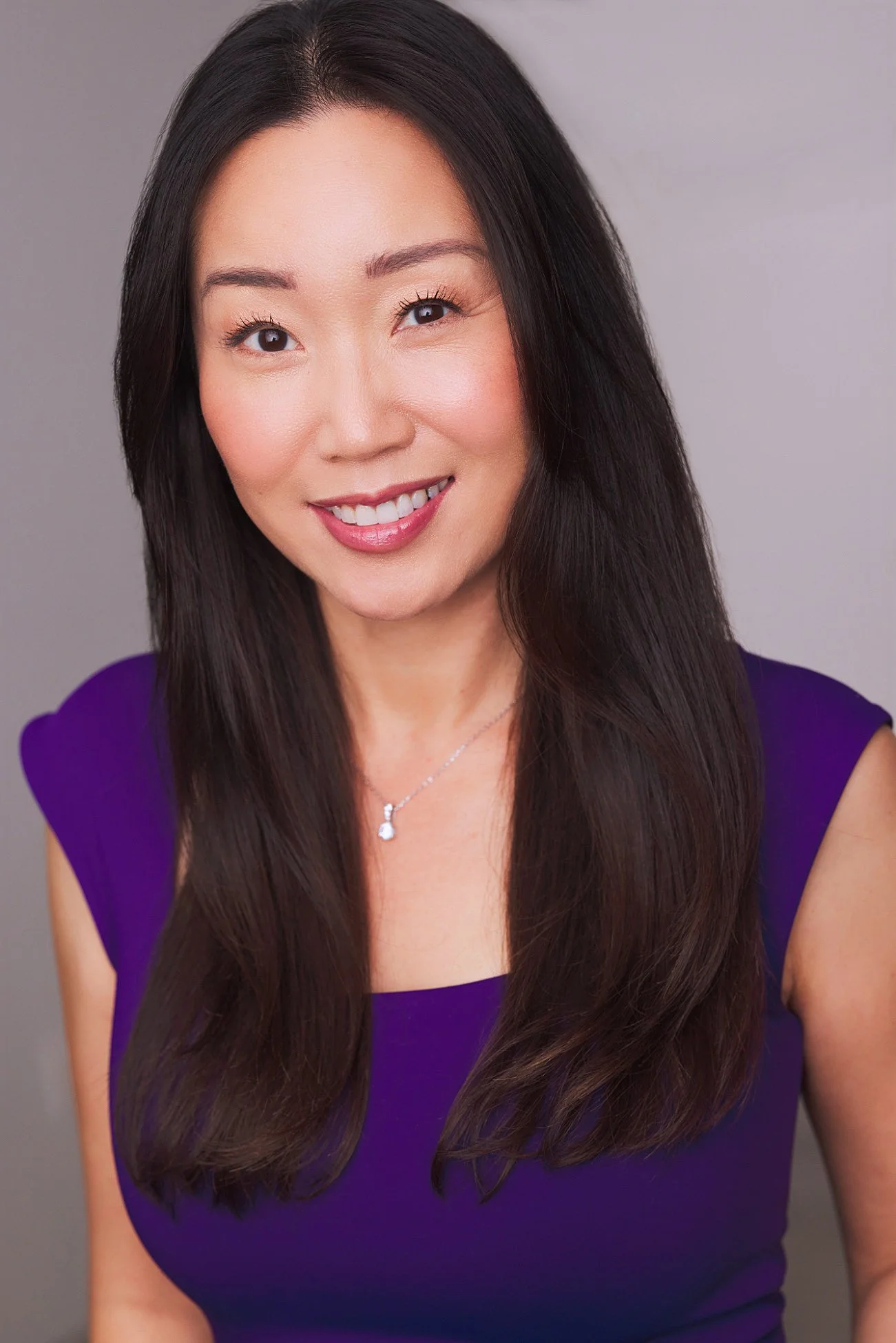 Portrait of a woman with long dark hair, smiling, wearing a purple top and a silver necklace with a teardrop-shaped pendant, against a plain gray background.