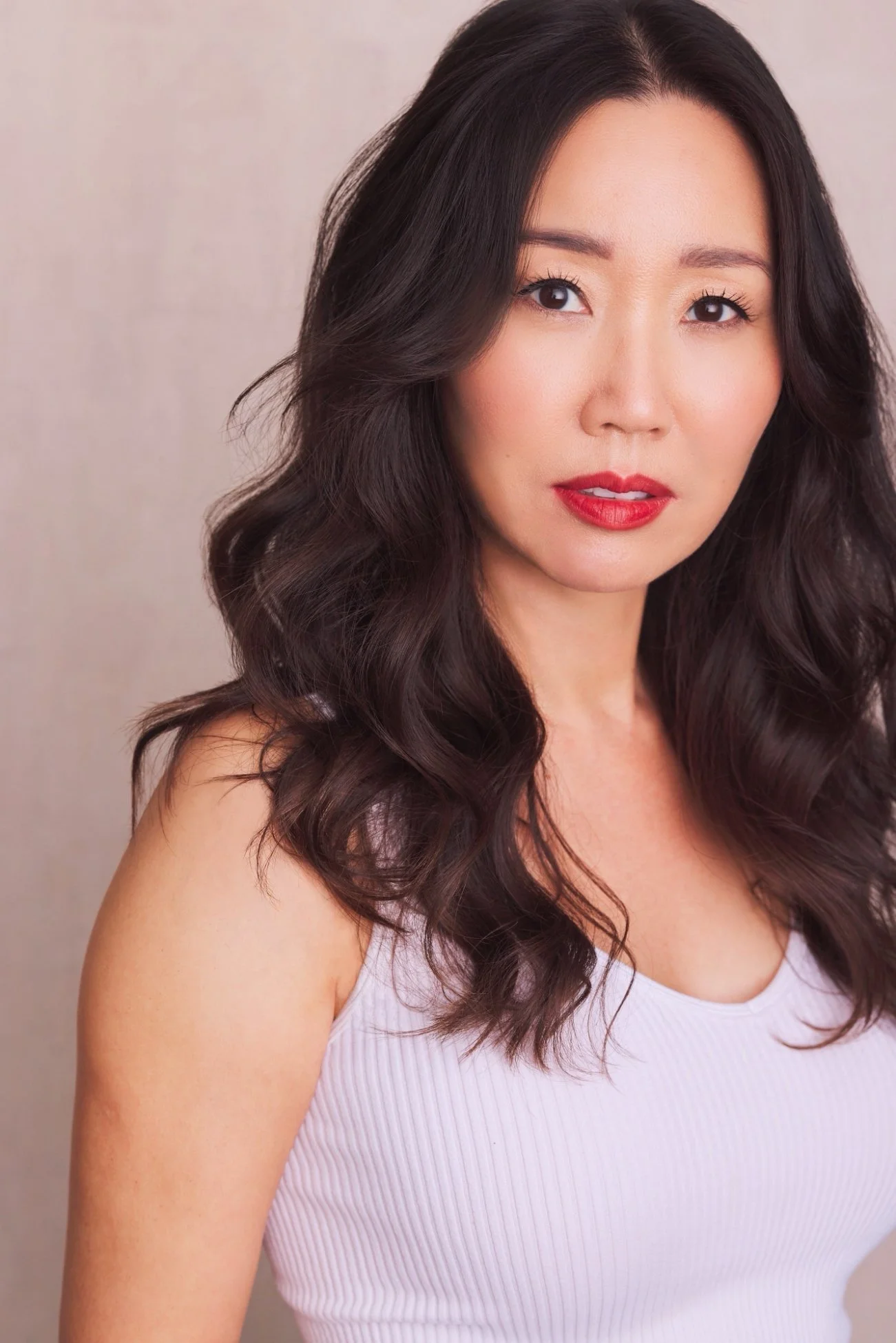 A woman with long, dark wavy hair wearing a white sleeveless top, looking at the camera with a neutral expression.