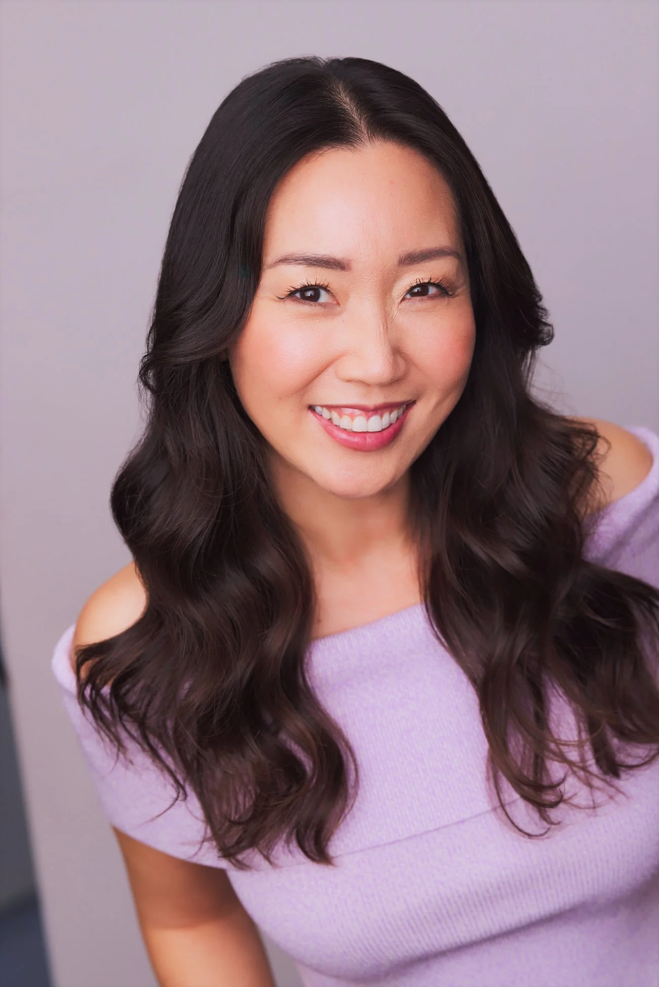 A woman with long, wavy dark hair wearing a lavender top, smiling at the camera.