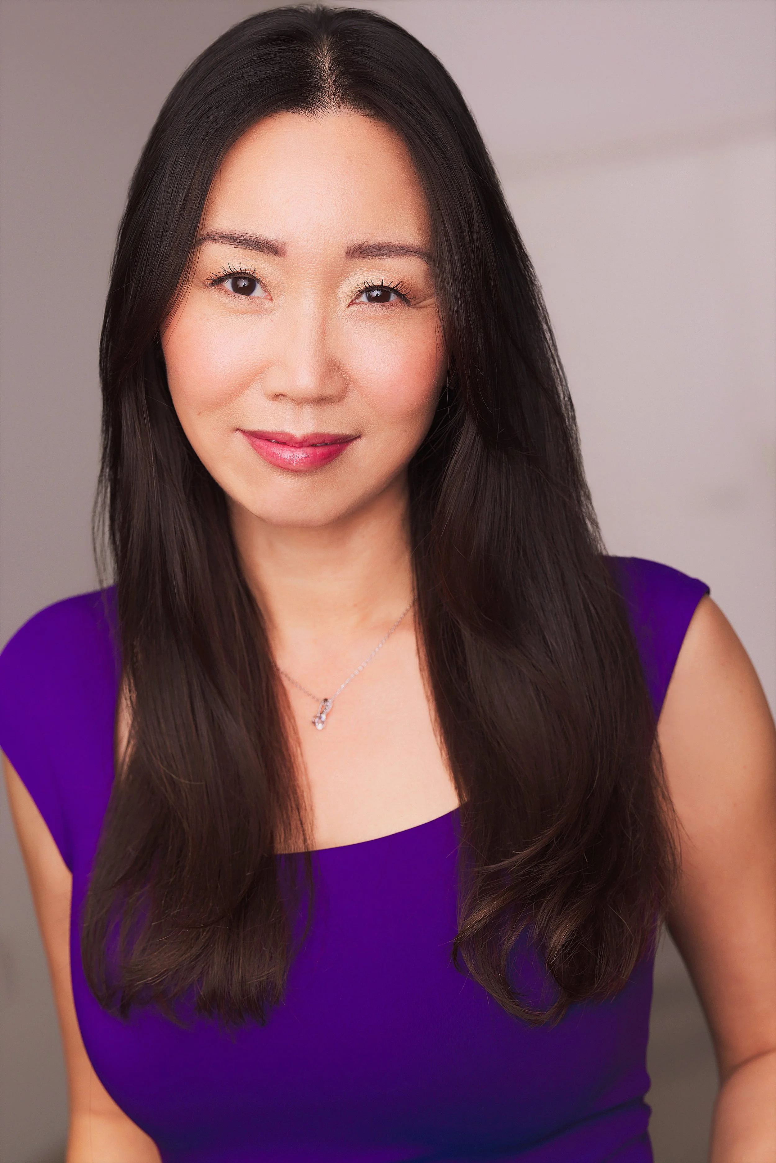 A woman with long dark hair wearing a purple top and a silver necklace, smiling gently at the camera.