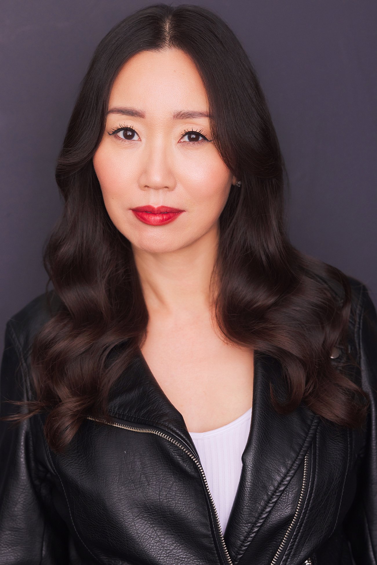 A woman with long wavy dark hair, wearing red lipstick, a white top, and a black leather jacket, posed against a dark background.