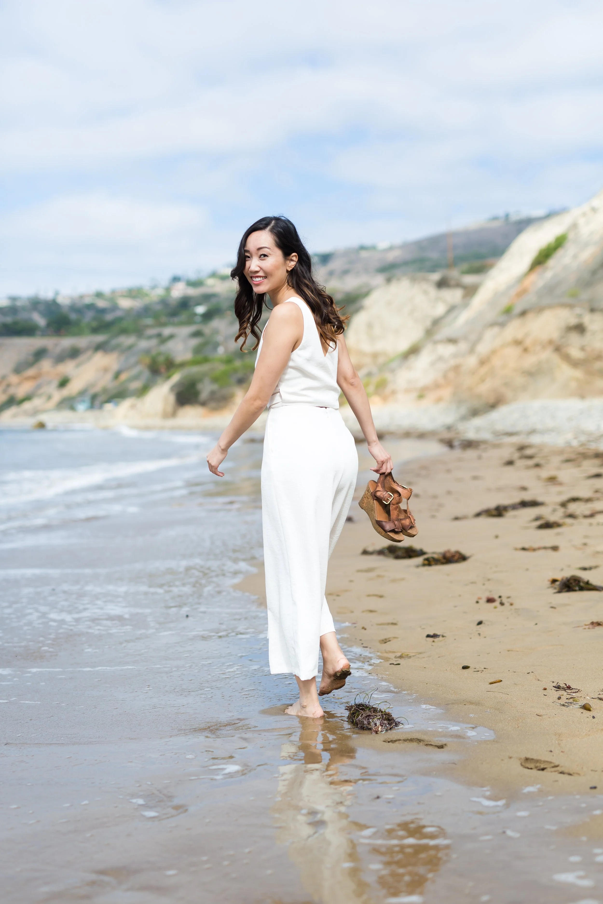 A woman in a white sleeveless jumpsuit walking barefoot along the shoreline of a beach, holding her brown sandals, with cliffs and a cloudy sky in the background.