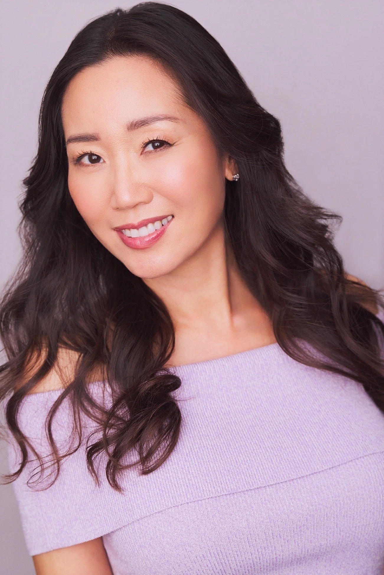 Portrait of a woman with long, wavy dark hair, smiling, wearing a light purple off-shoulder top, against a neutral background.