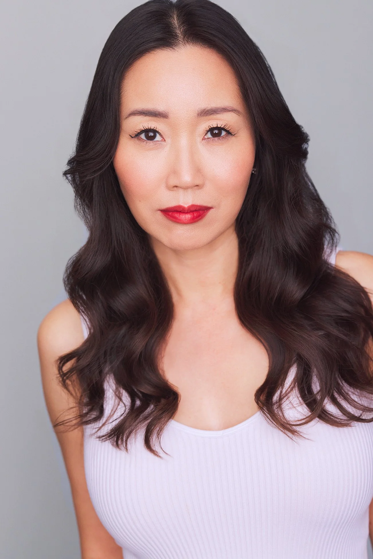 A woman with long dark wavy hair and red lipstick looking directly at the camera, wearing a white sleeveless top, against a neutral background.