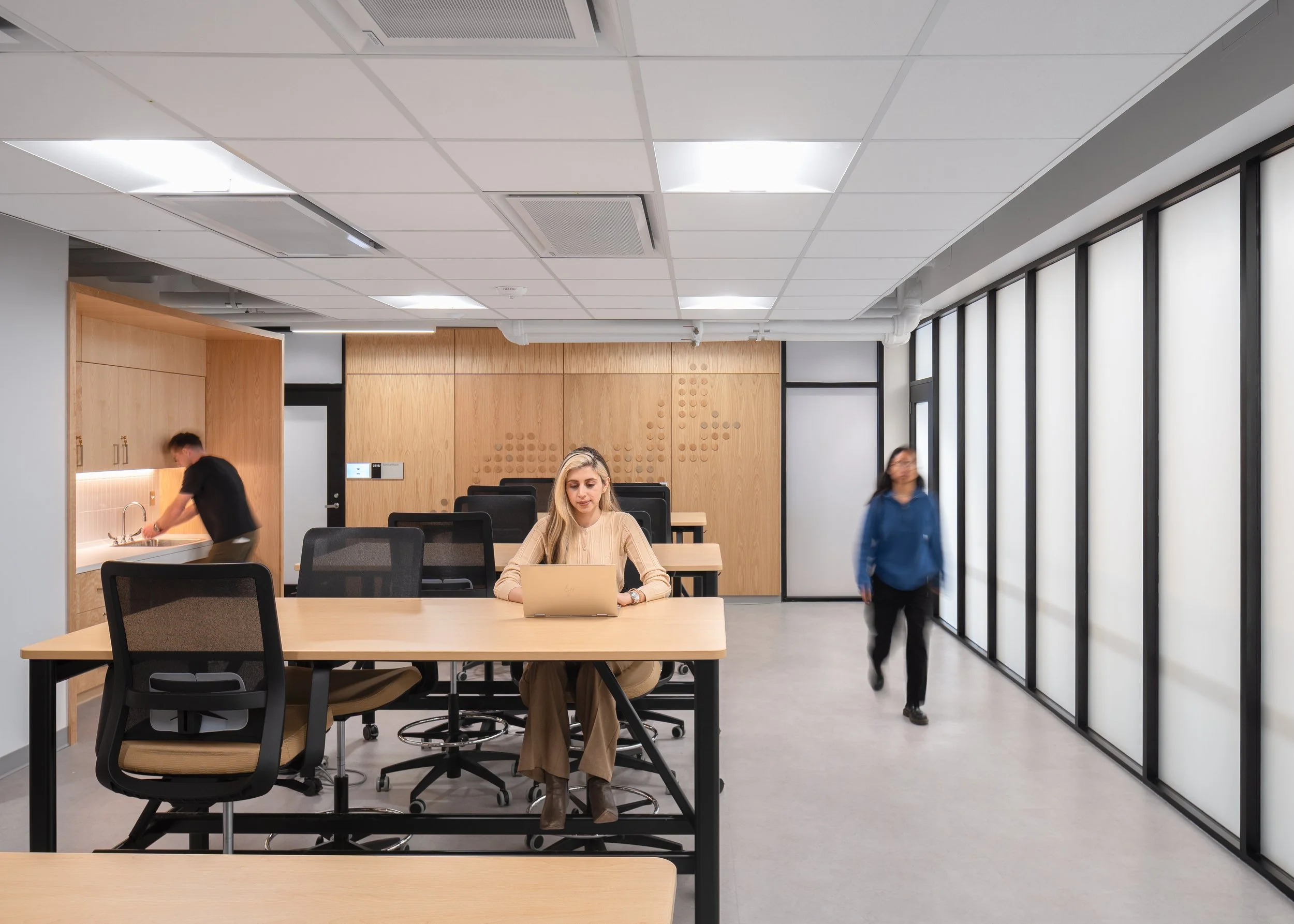 A modern office with a woman sitting at a table working on a computer, two people in the background walking, large windows on the right, and a kitchenette area on the left.