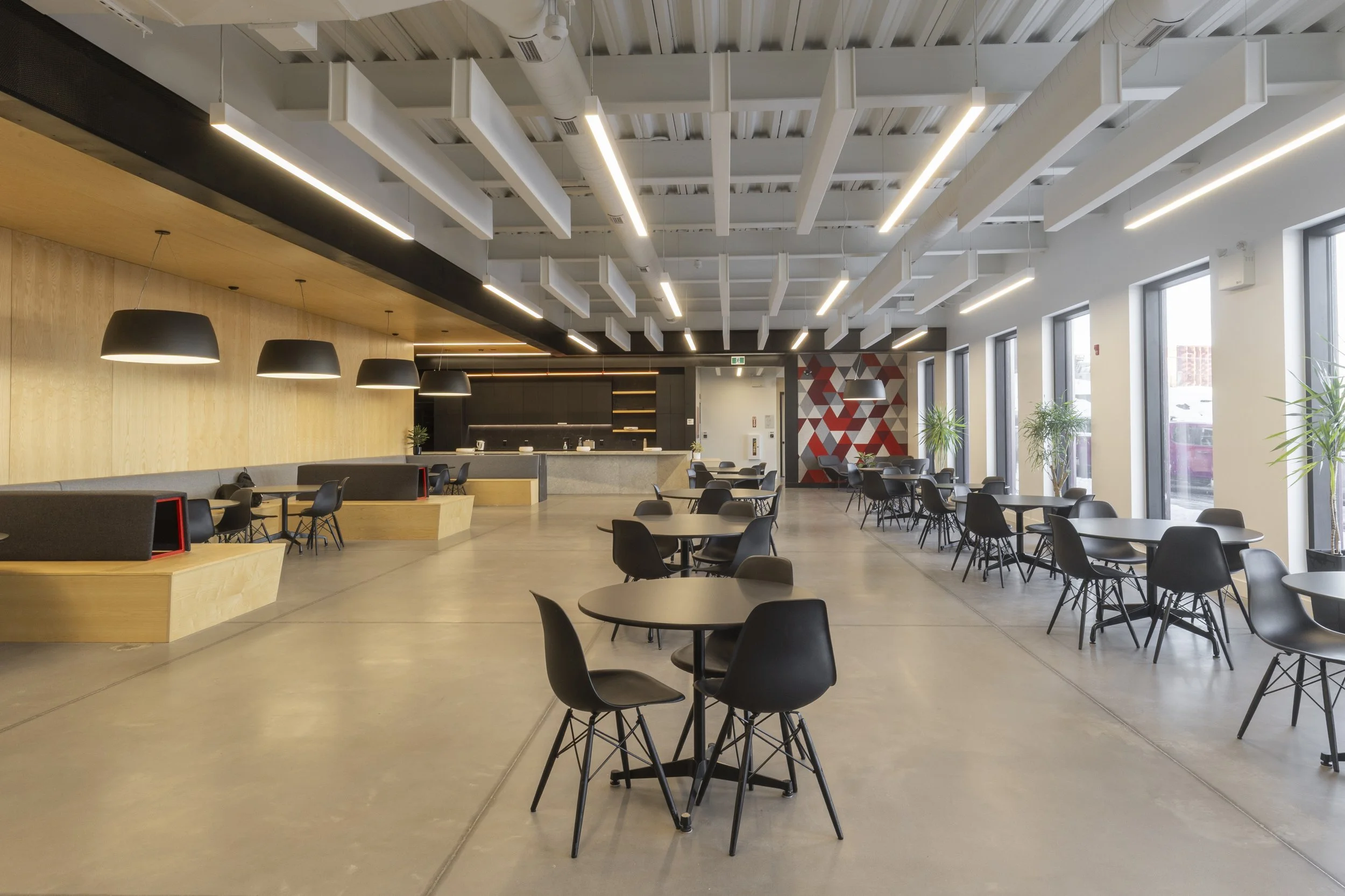 Interior of a modern, spacious cafeteria or dining area with black round tables, black chairs, large windows, greenery, wood-paneled walls, and a ceiling with white beams and lighting fixtures.