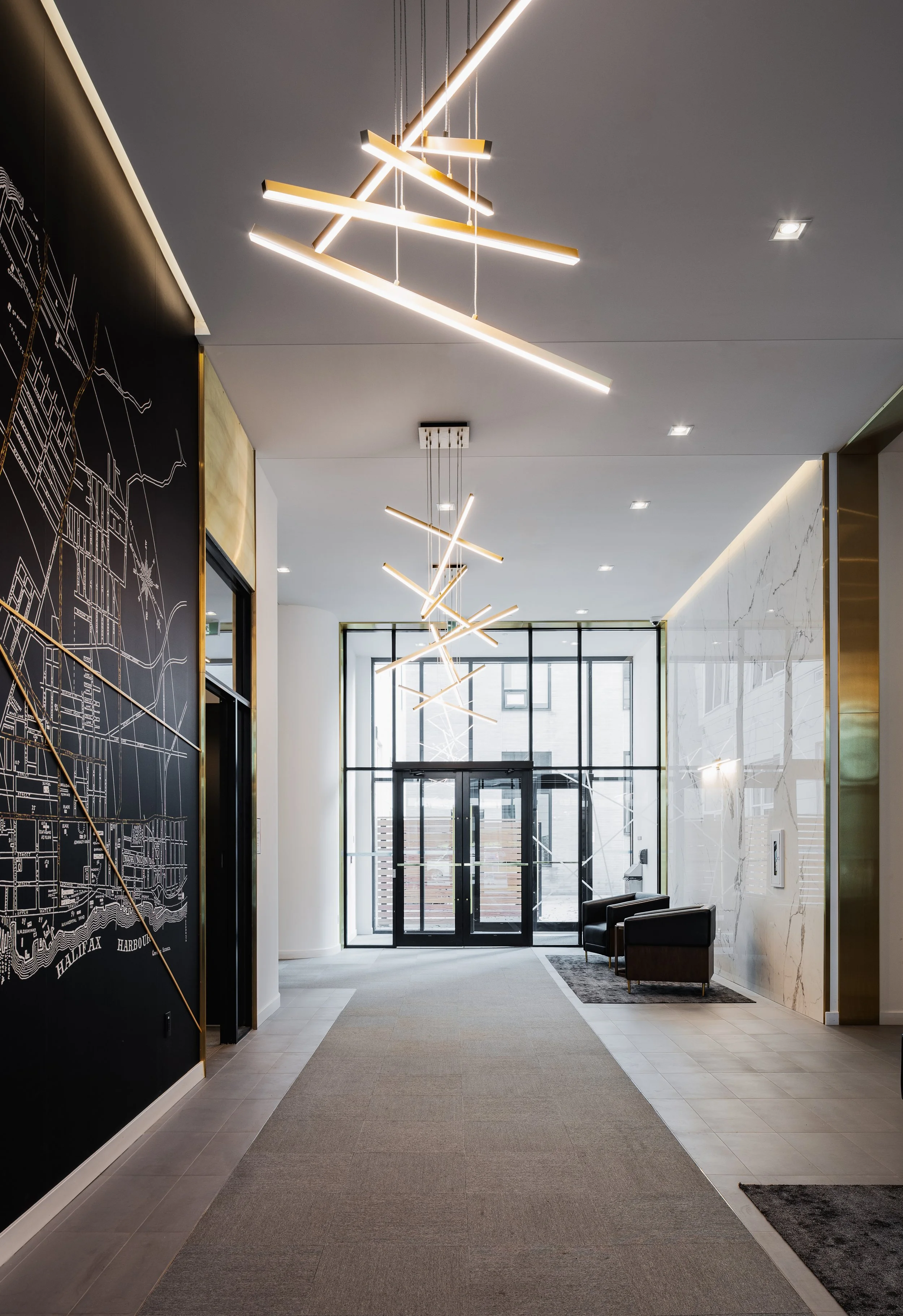 Modern lobby with black wall decorated with a white city map, seating area with two black chairs, a gray rug, large glass front entrance, and contemporary linear and geometric ceiling lights.