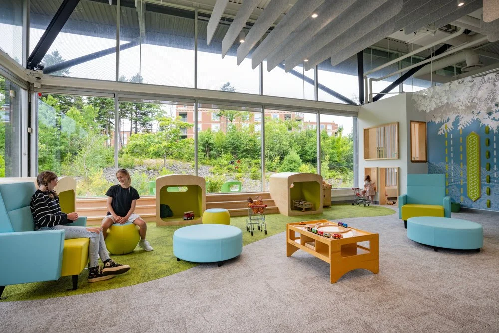 Children playing and sitting in a brightly lit, modern indoor playroom with large windows, colorful furniture, and outdoor greenery visible.