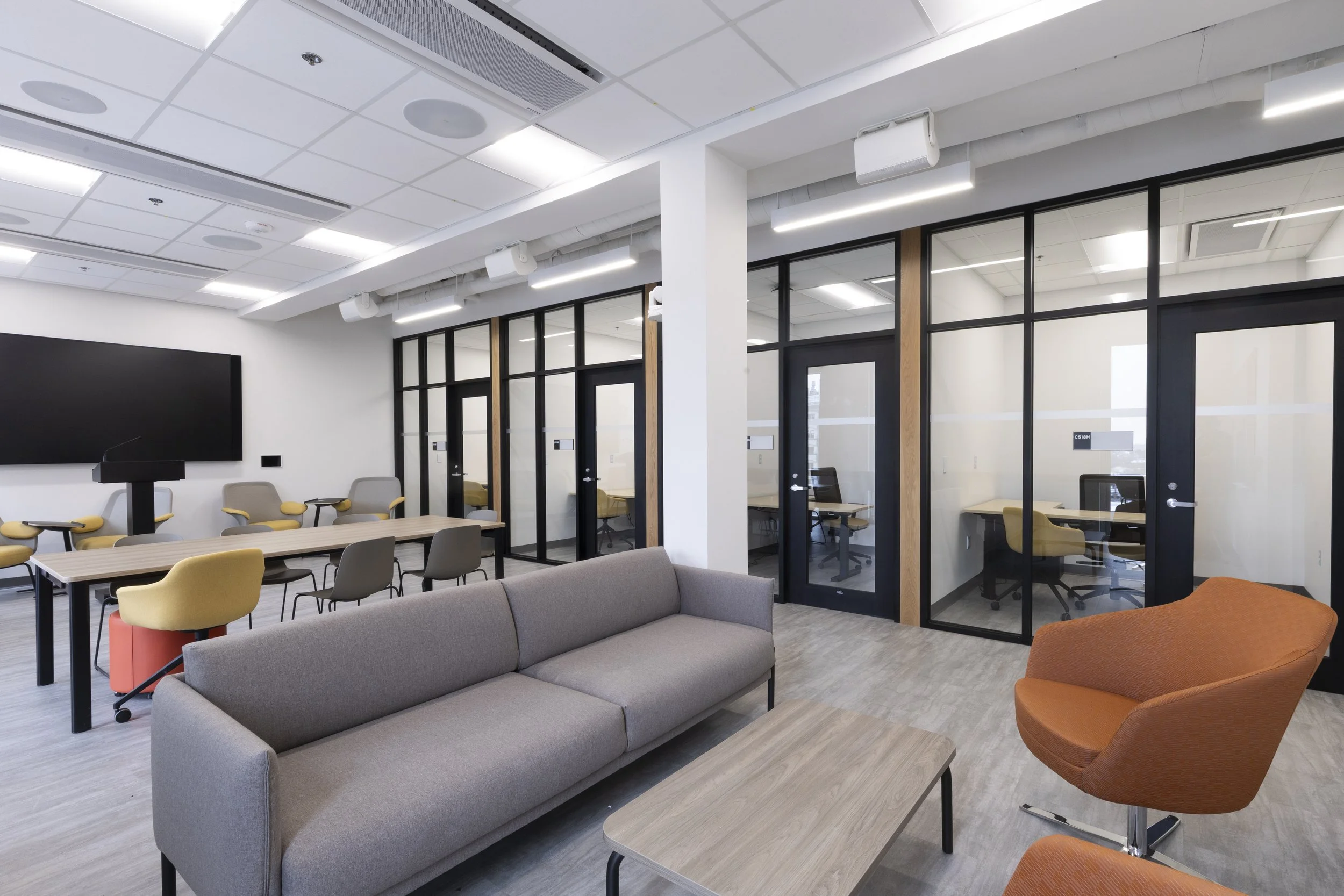 Modern office conference room with a ceiling-mounted flat screen TV, rows of chairs, glass partitioned enclosed workspaces, and a mix of gray, orange, and yellow furniture.