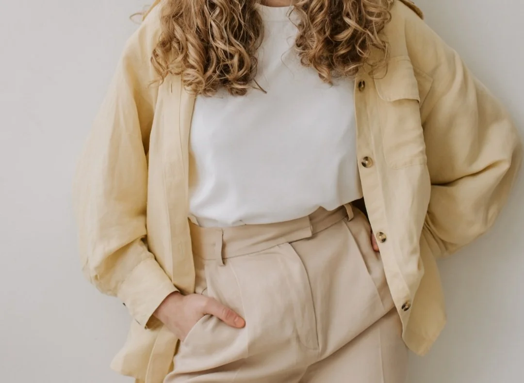 A person with curly hair wearing a beige jacket over a white t-shirt and beige pants, standing against a light-colored wall.