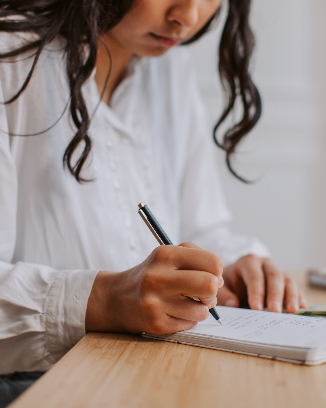 A woman in a white blouse writing in a notebook with a black pen on a wooden desk.