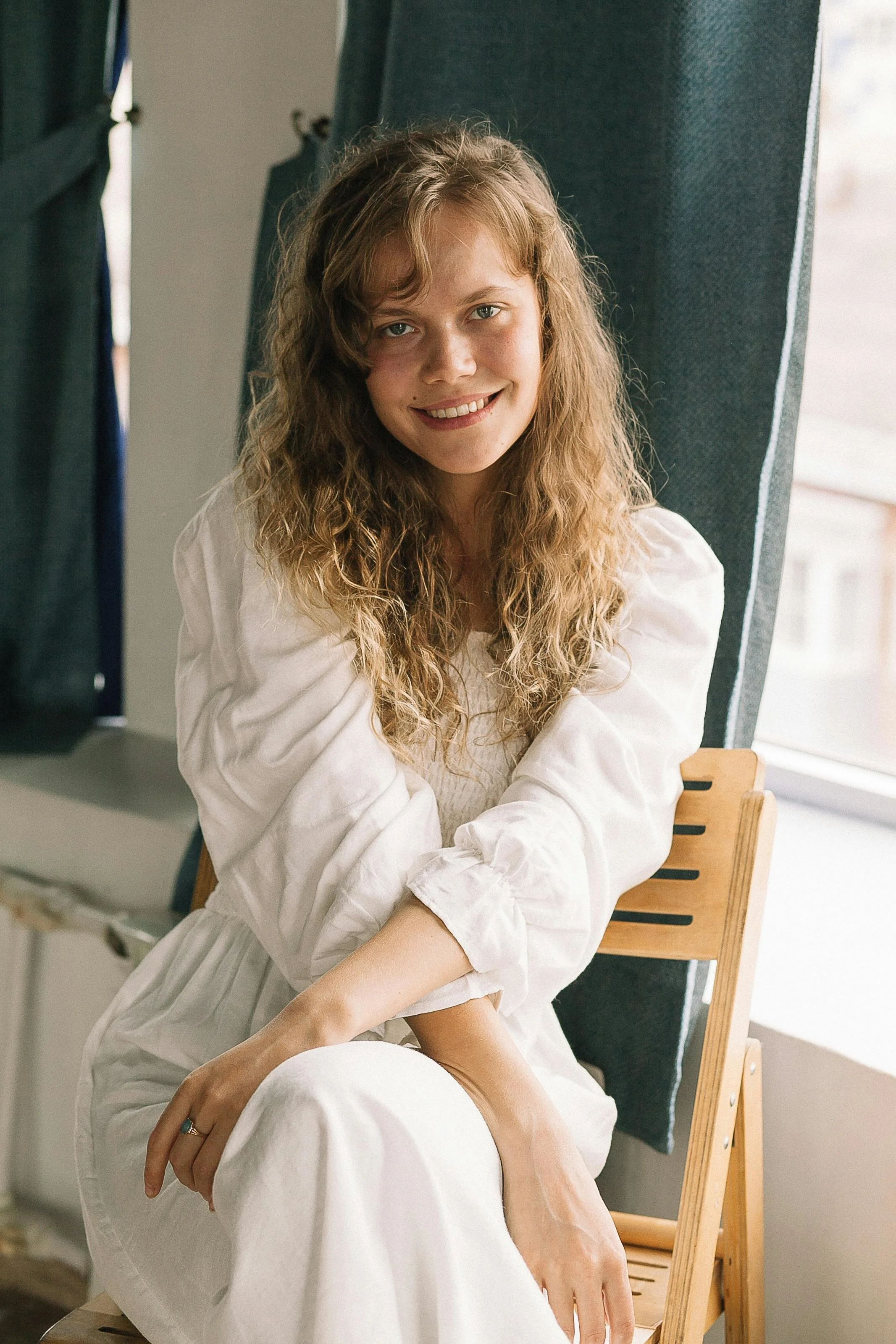 A young woman with curly blonde hair sitting on a wooden chair in front of a window with dark curtains, smiling and wearing a white dress.