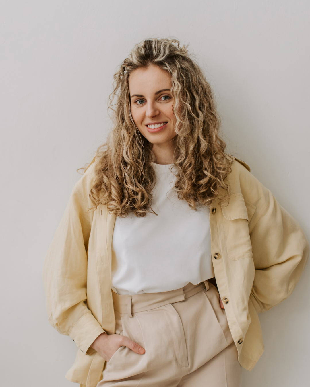 A young woman with curly blonde hair wearing a light beige jacket over a white T-shirt and beige pants, standing against a plain white wall, smiling and posing casually.
