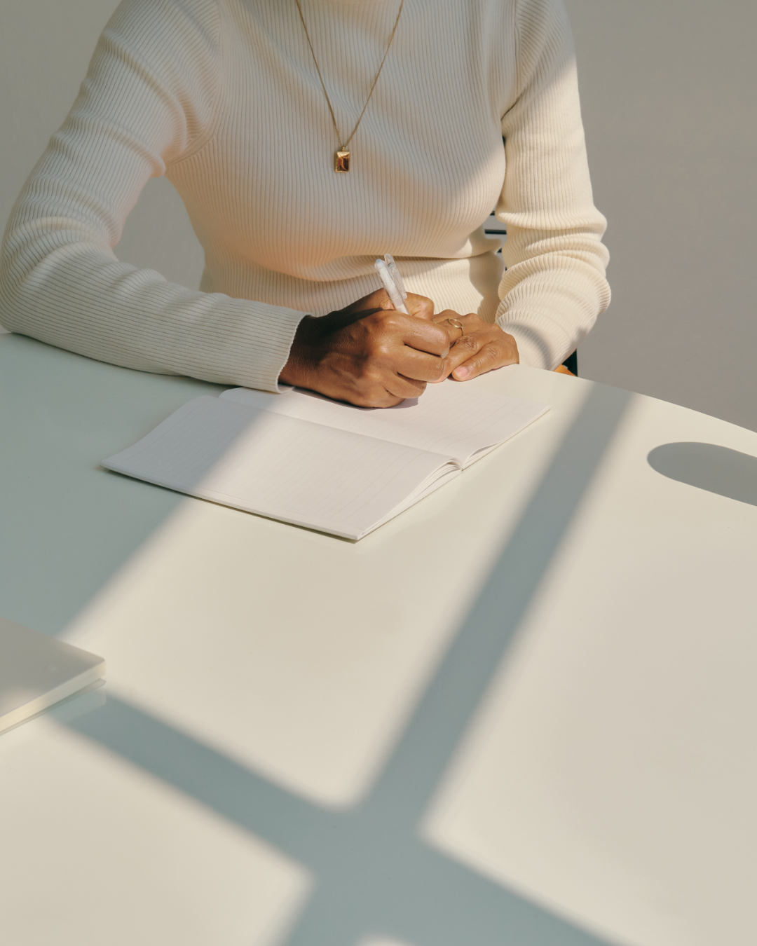 A person writing in a notebook at a white table with sunlight casting shadows.