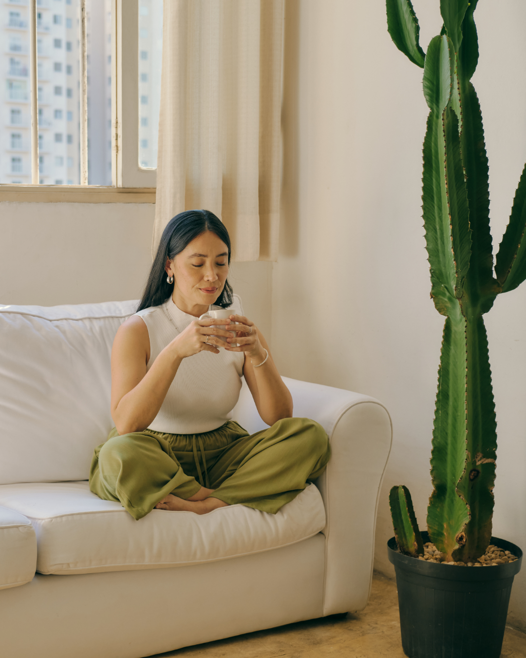 A woman sitting cross-legged on a white sofa, holding a mug, in a room with a large cactus plant and windows with city buildings outside.