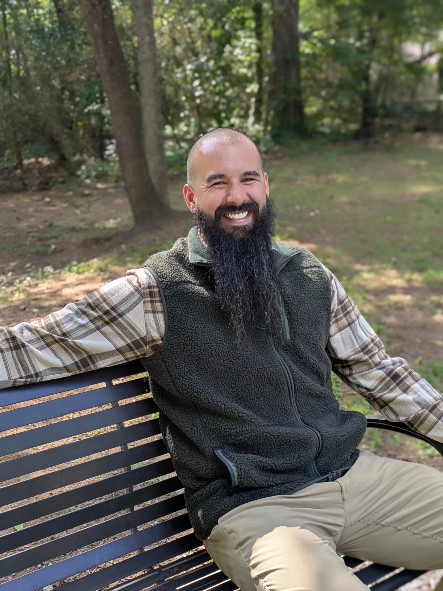 A bearded man smiling and sitting on a park bench during daytime, wearing a plaid shirt and a fleece vest, with trees and greenery in the background.