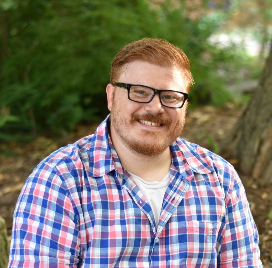 A smiling man with glasses and red hair outdoors surrounded by green foliage.