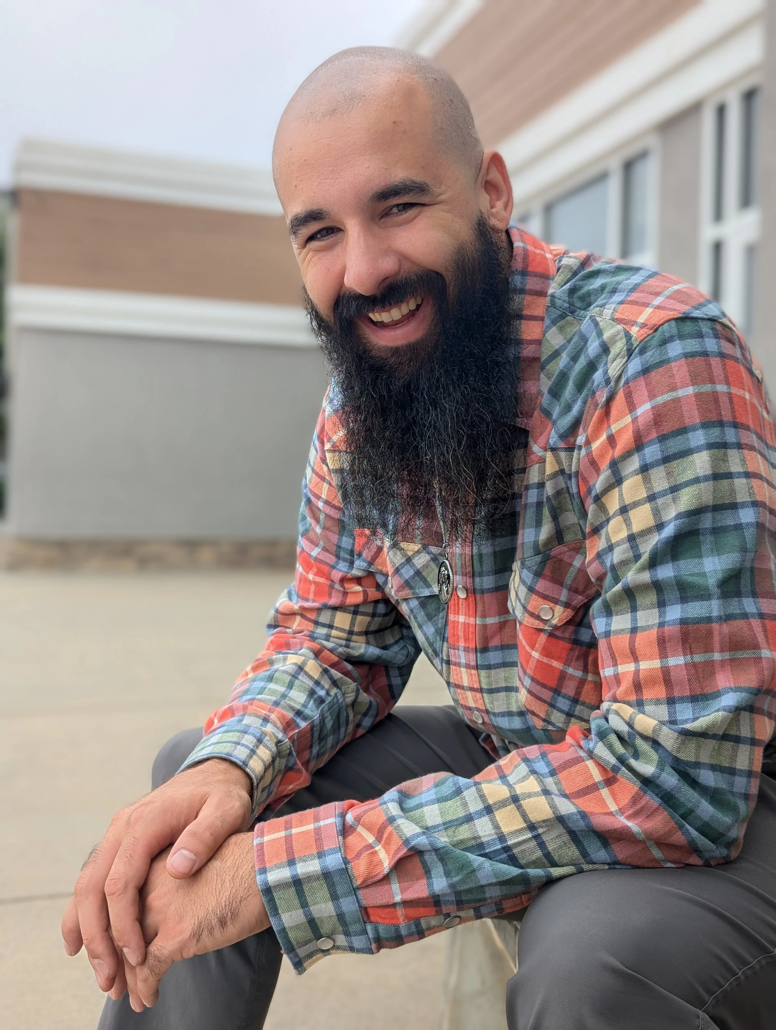 A man with a large beard, smiling, wearing a colorful plaid shirt sitting outdoors.