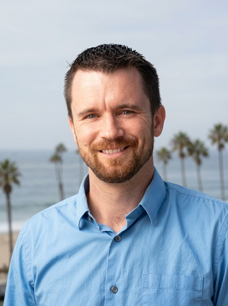 A man with a beard and mustache wearing a light blue collared shirt, smiling with palm trees and the ocean in the background.