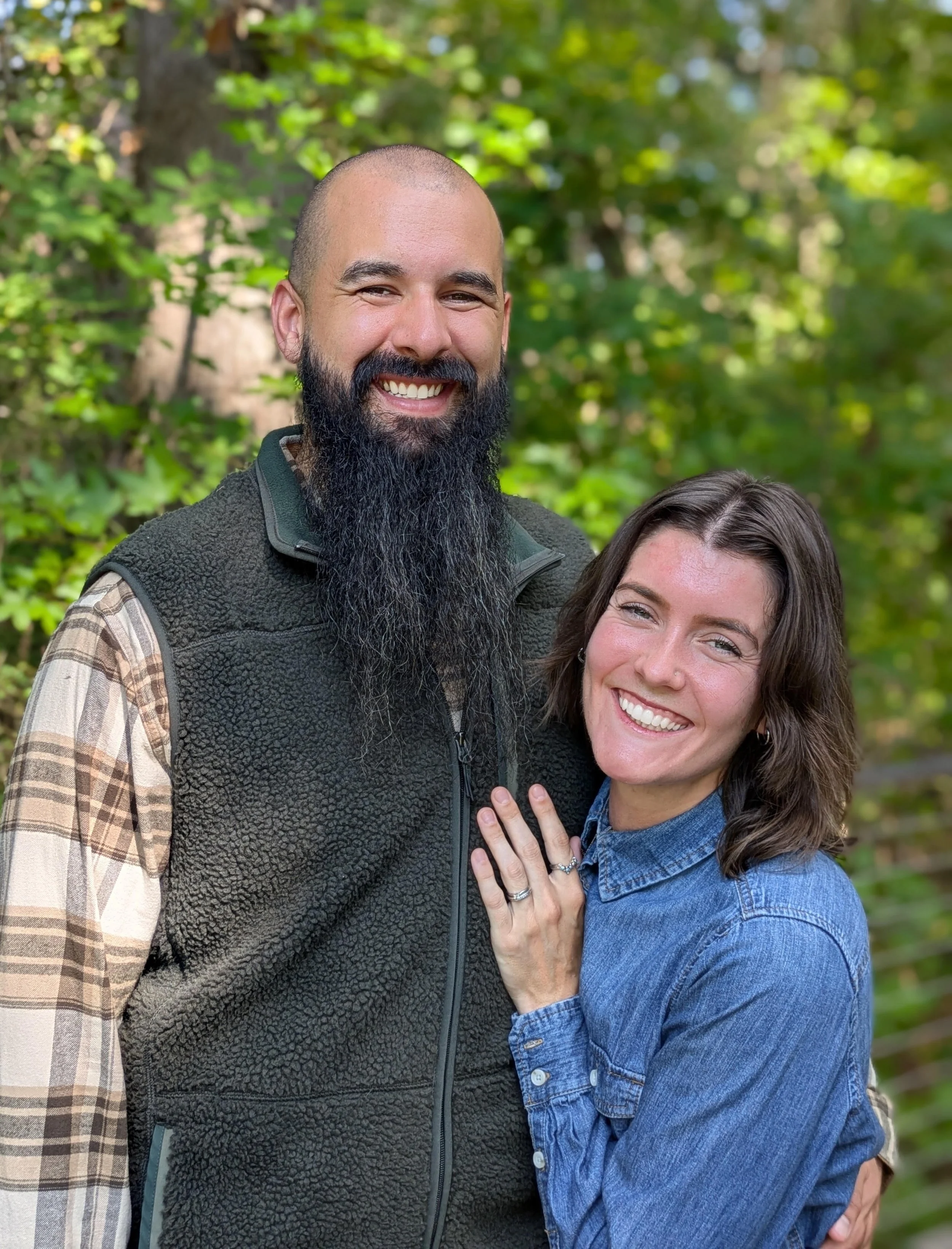 A smiling man with a long beard wearing a plaid shirt and a dark vest standing outdoors with a woman with dark hair and a ring on her finger, in front of green trees.
