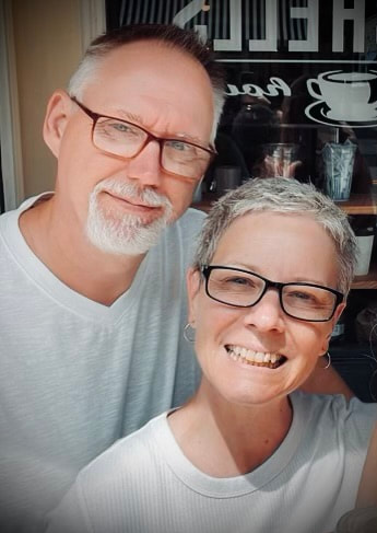 A middle-aged man and woman smiling for a selfie outside a café, both wearing glasses and white shirts.