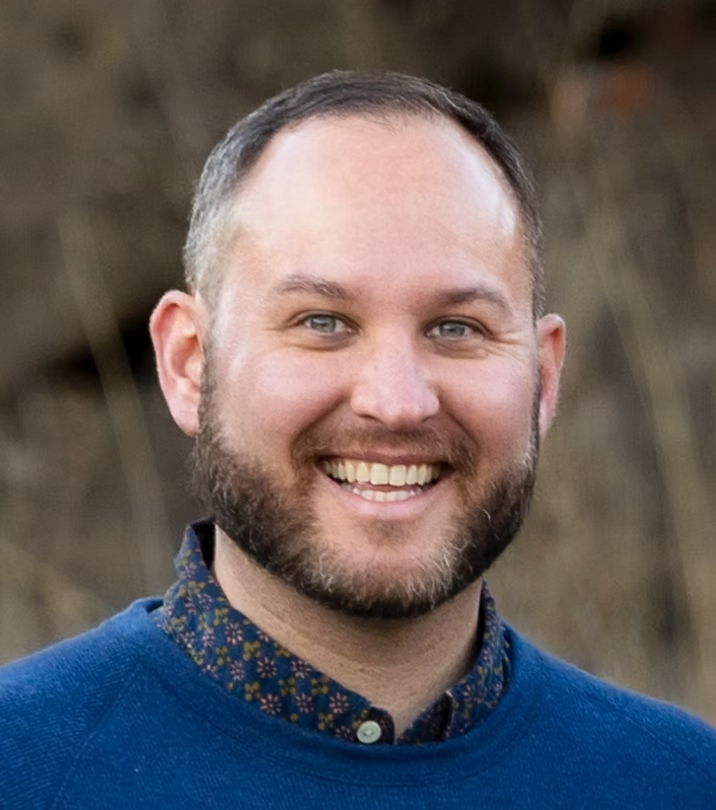 A smiling man with short hair, a beard, and blue eyes outdoors with blurred natural background.