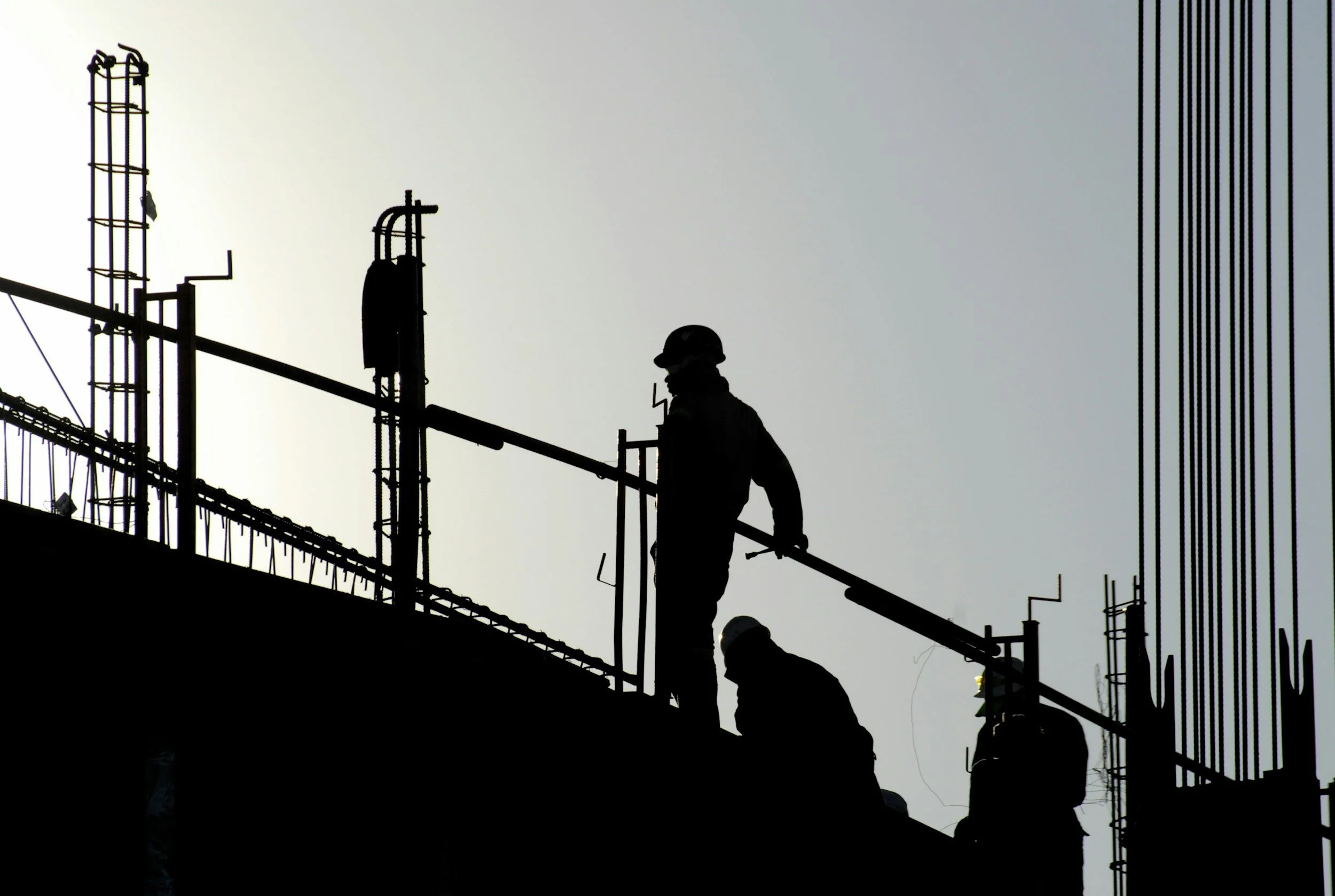Silhouettes of construction workers working on a building under construction, with visible scaffolding and equipment against a gray sky.