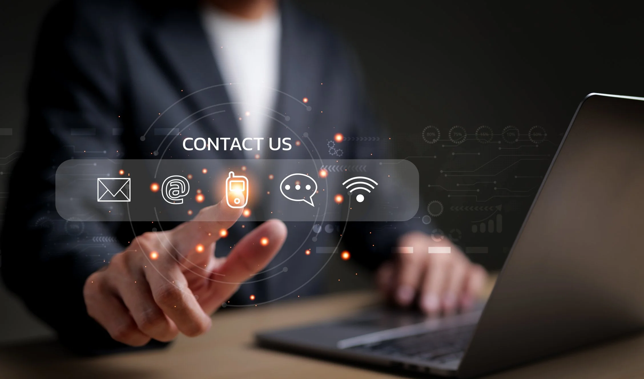 A person in a dark suit interacts with a holographic contact menu featuring icons for email, at symbol, mobile phone, chat bubble, and Wi-Fi, with a laptop on the table.