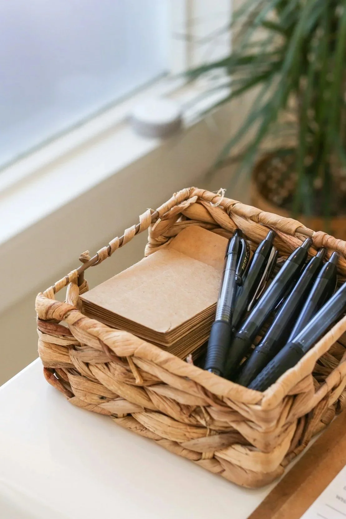 Wicker basket on a white table holding blank notepads and black pens, with a plant and window in the background.