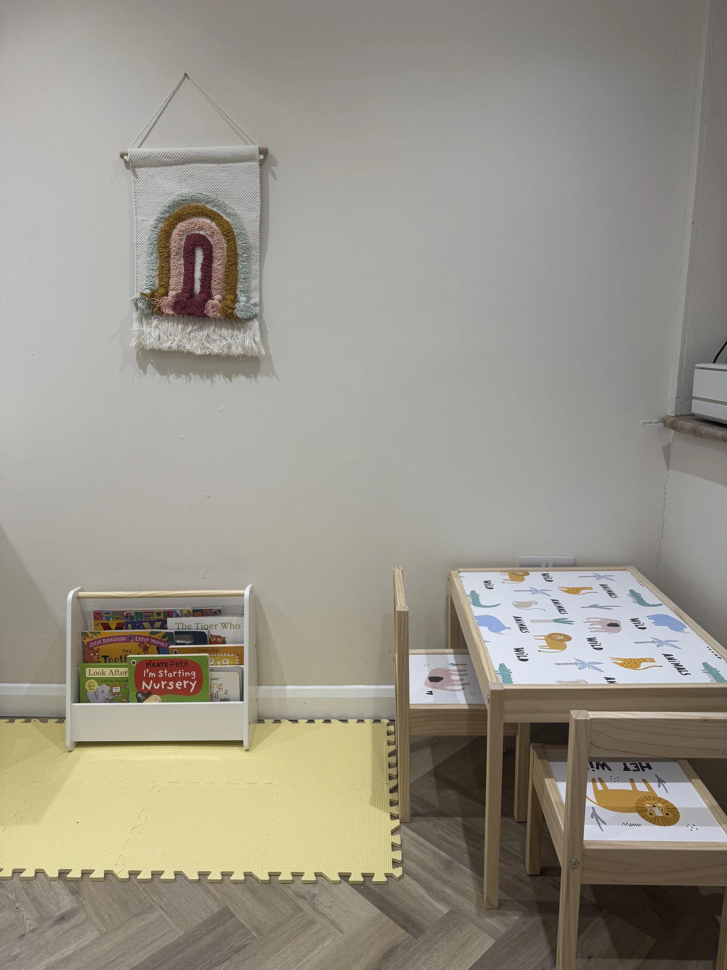 Children's reading and play area with a white bookshelf holding children's books, a small wooden table with animal-themed top, and a wooden child's chair, on a yellow foam play mat, against a plain wall with a colorful rainbow wall hanging.