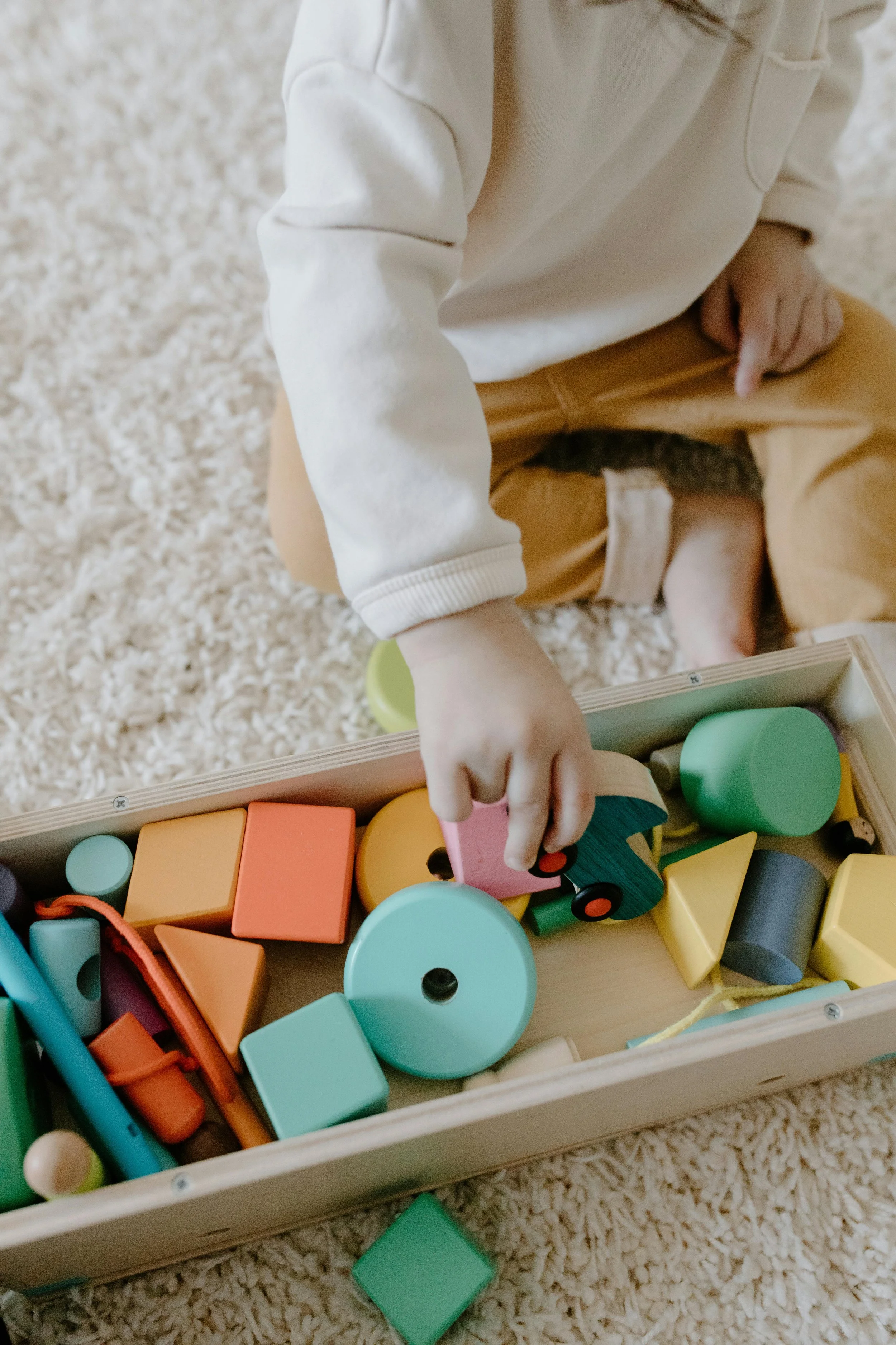 Child playing with colorful wooden building blocks in a wooden box, sitting on a plush beige carpet.