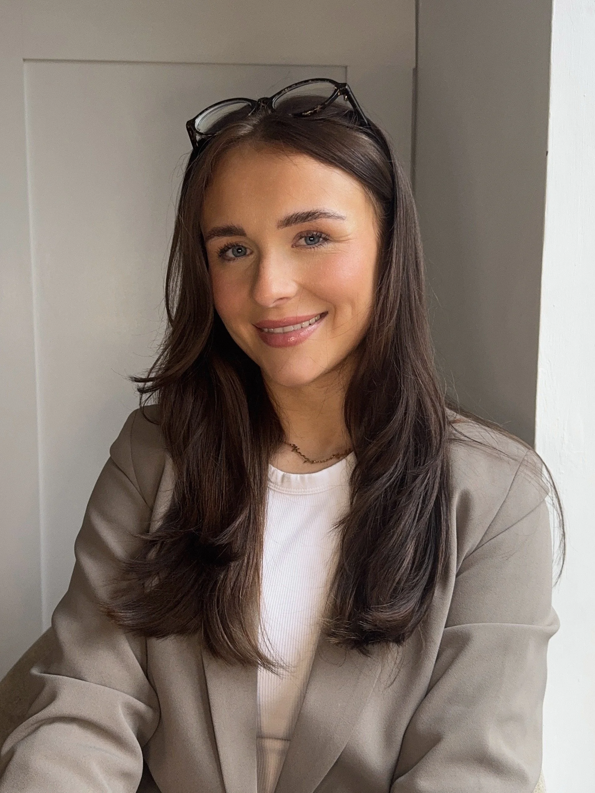 A young woman with long brown hair, blue eyes, and light skin, smiling with her teeth visible. She is wearing glasses on her head, a beige blazer, a white top, and a small necklace. She is seated indoors near a white wall and door.