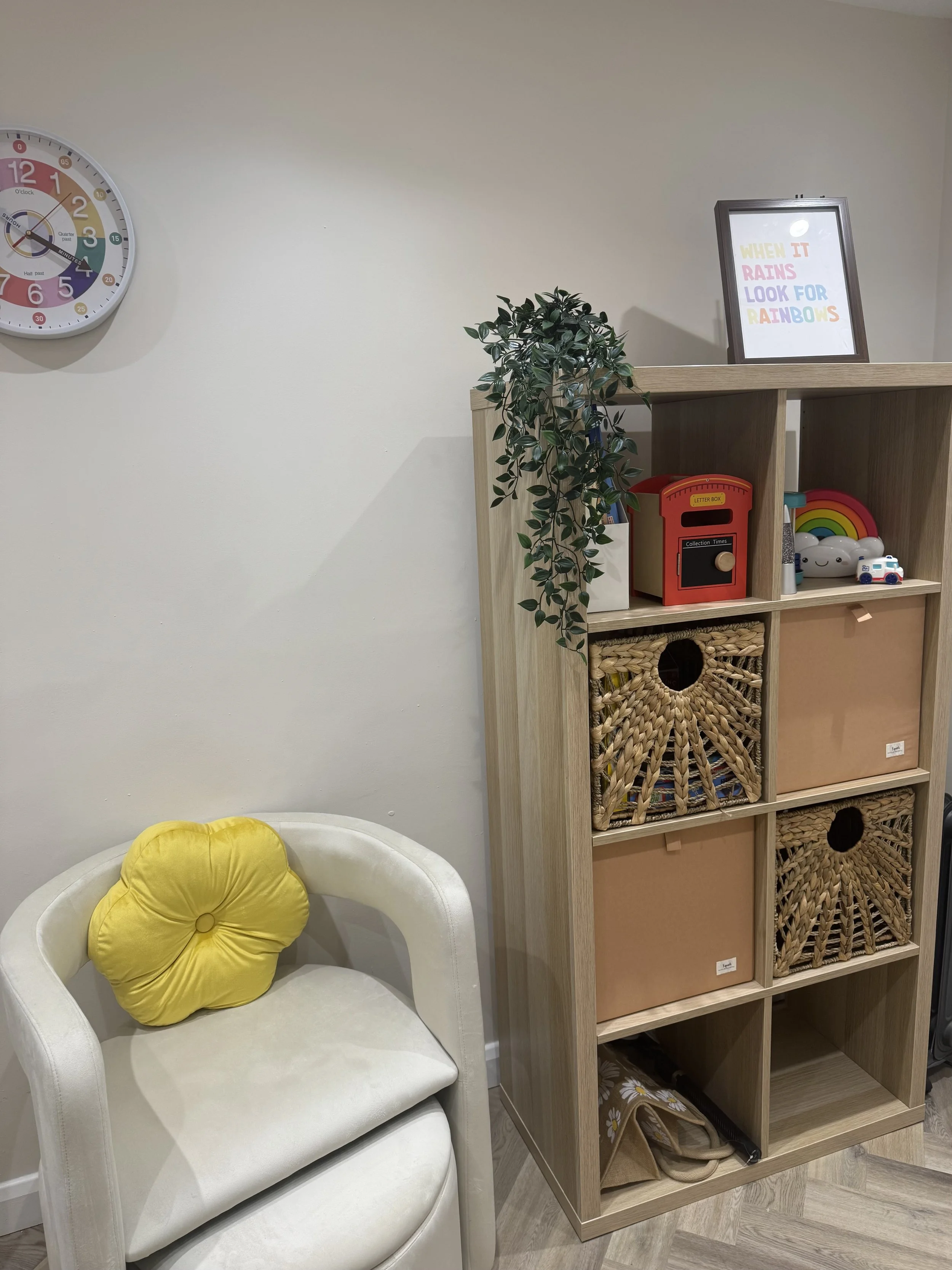 A white armchair with a yellow flower-shaped cushion, a wooden shelf with decorative storage bins and toys, a potted plant, and a colorful motivational sign on top of the shelf, with a wall clock in the background.