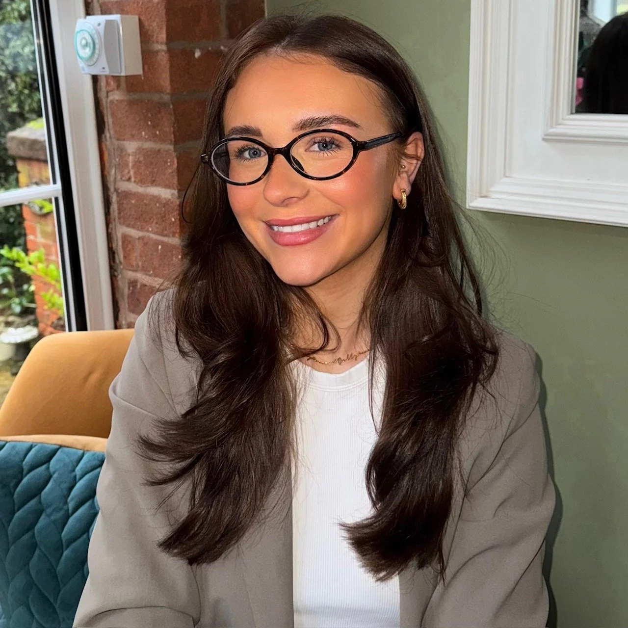 A young woman with long dark hair, wearing glasses, a grey blazer, and a white top, smiling while seated indoors.