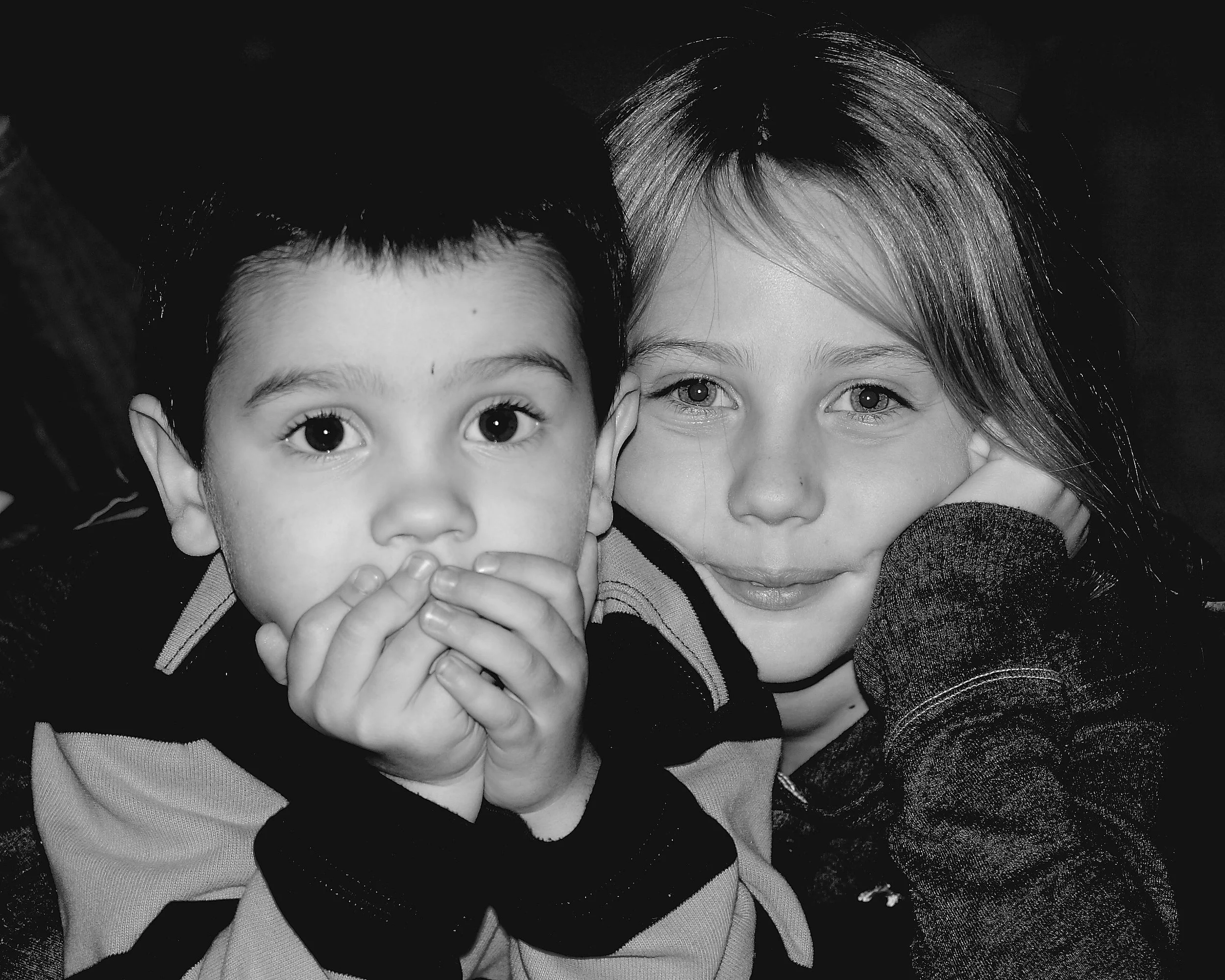 Black and white photo of a young boy with short dark hair and a young girl with long light hair, both resting their heads on their hands and looking into the camera.