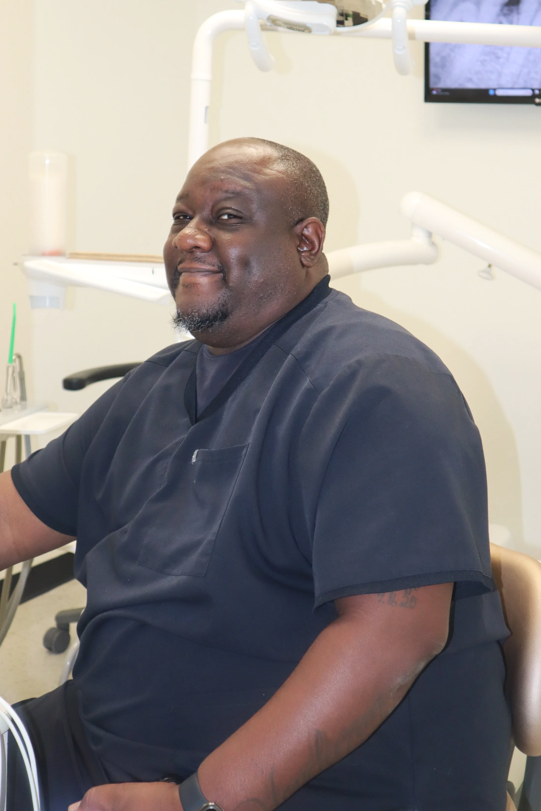 A smiling man in black scrubs sitting in a dental or medical office, with dental equipment and a monitor in the background.