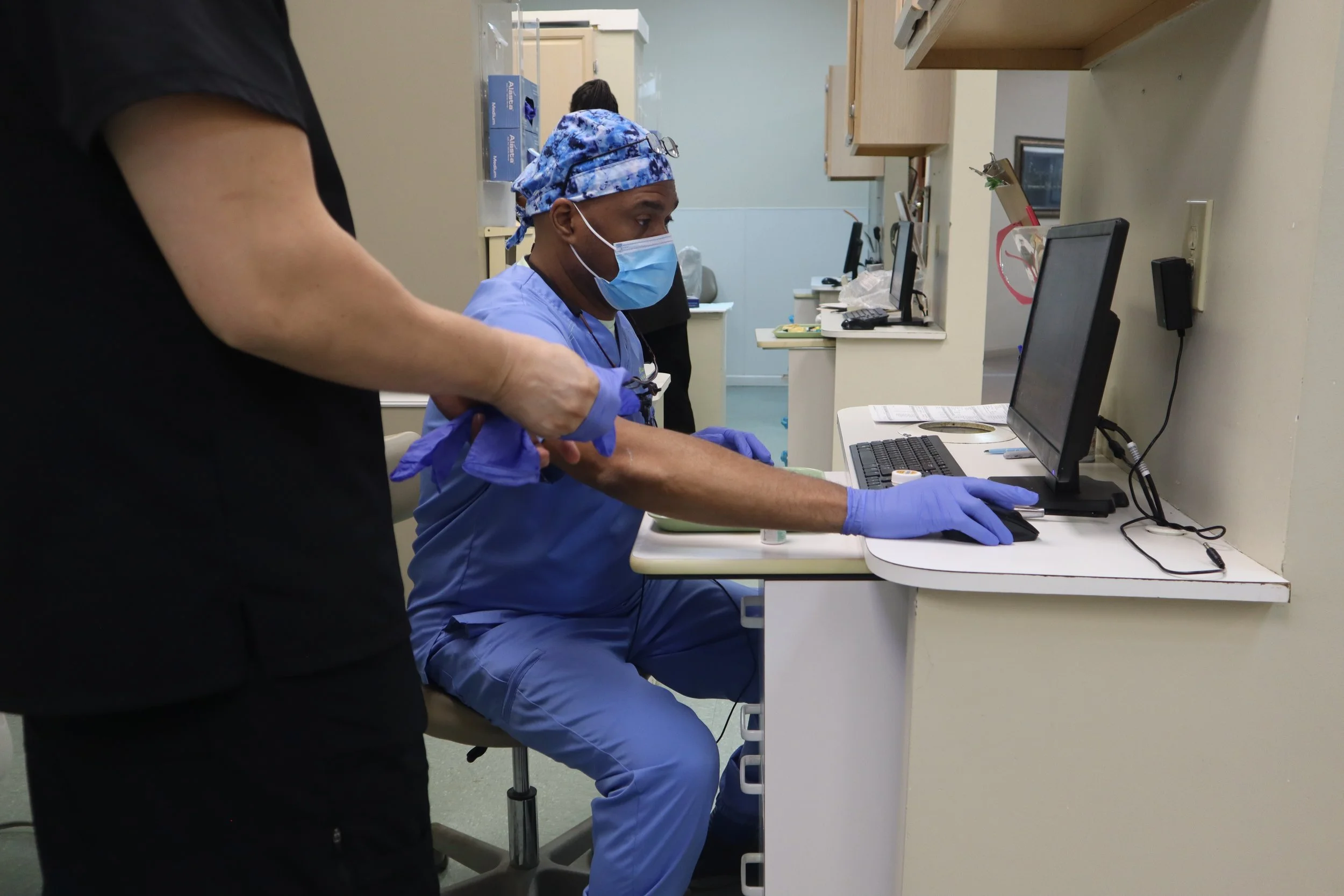 A medical professional in scrubs, wearing a face mask and gloves, sitting at a desk with a computer, with another person standing beside him, in a clinical setting.
