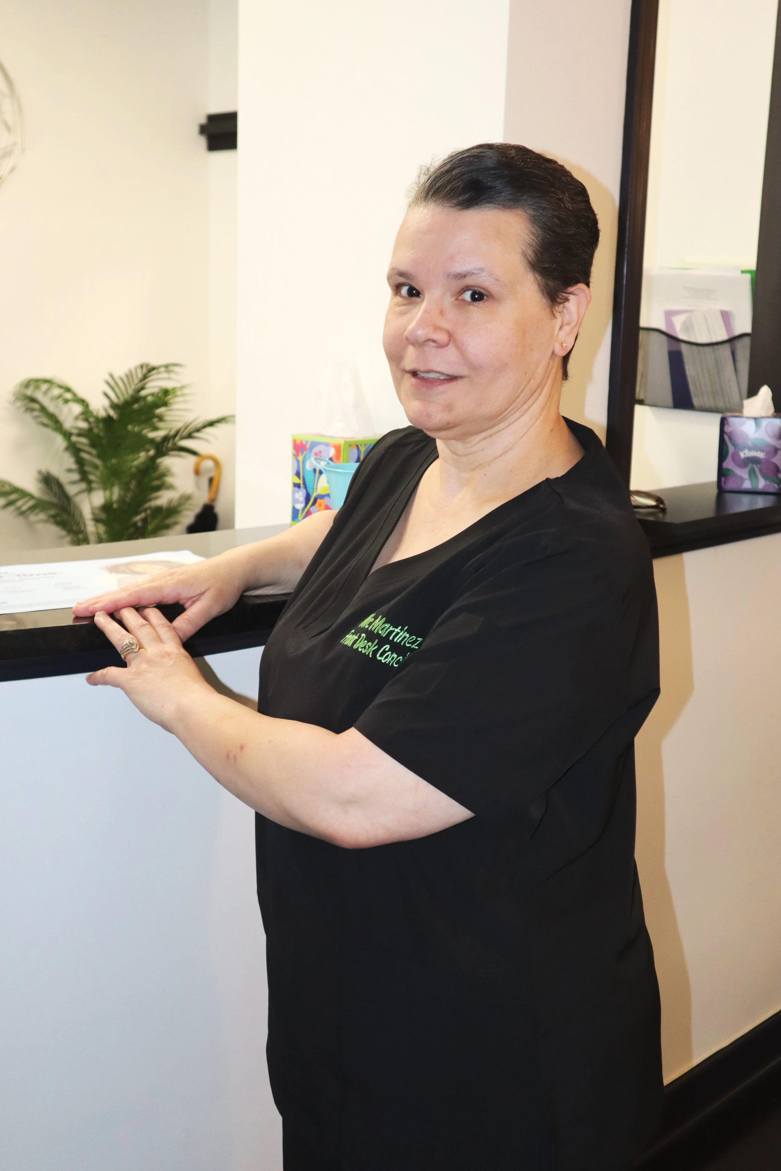 A woman in black scrubs leaning on a reception desk, smiling slightly at the camera, in a medical or dental office setting.