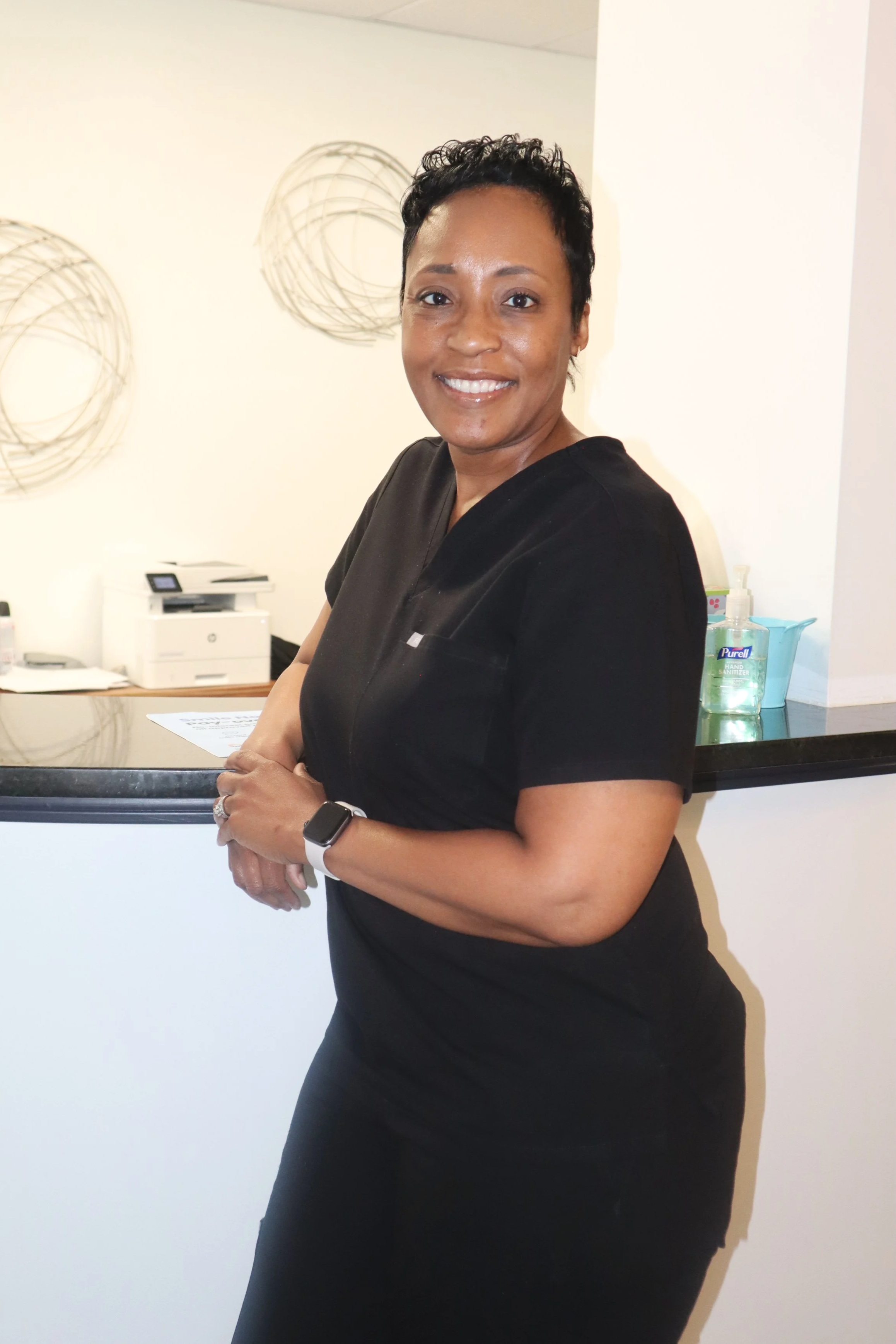 Smiling woman in black scrubs standing at a reception counter.