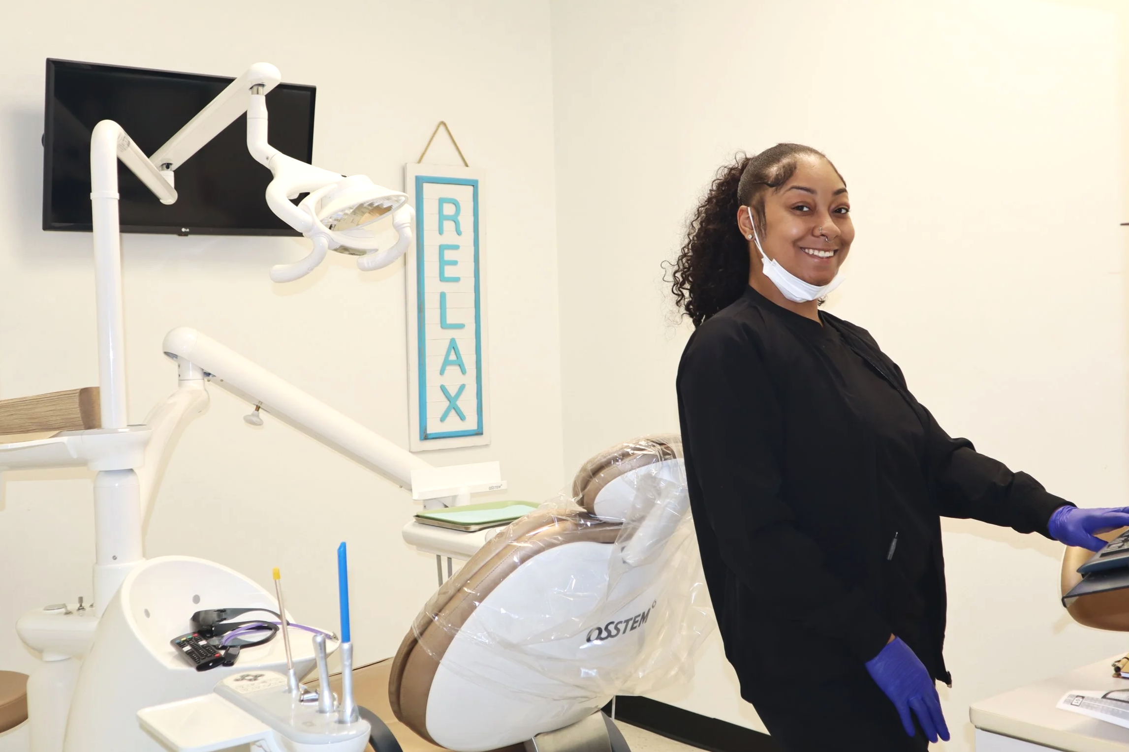 A woman in a dental office wearing a black scrub top, blue gloves, and a face mask pulled down, smiling at the camera. Dental equipment and a "RELAX" sign are visible in the background.