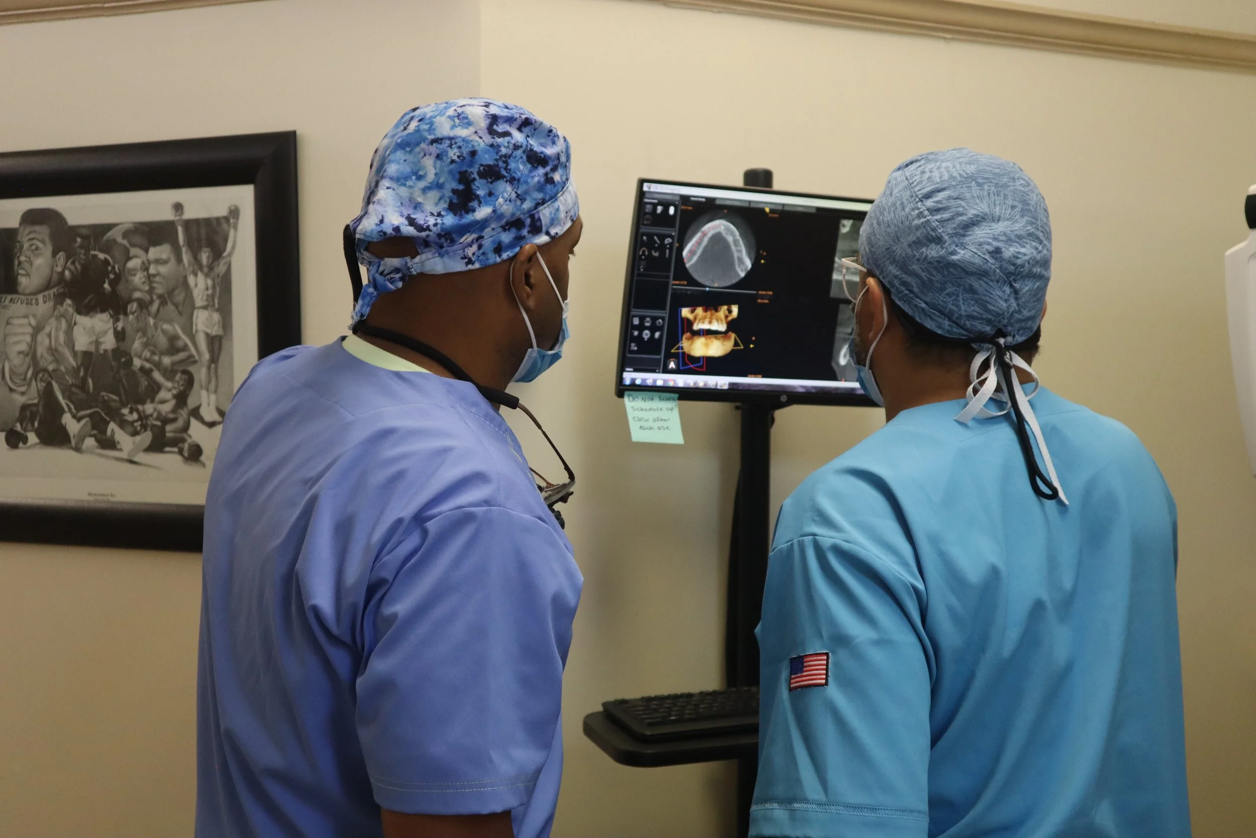 Two healthcare professionals wearing scrubs, masks, and caps are looking at a computer screen displaying dental X-rays or scans in a medical office.