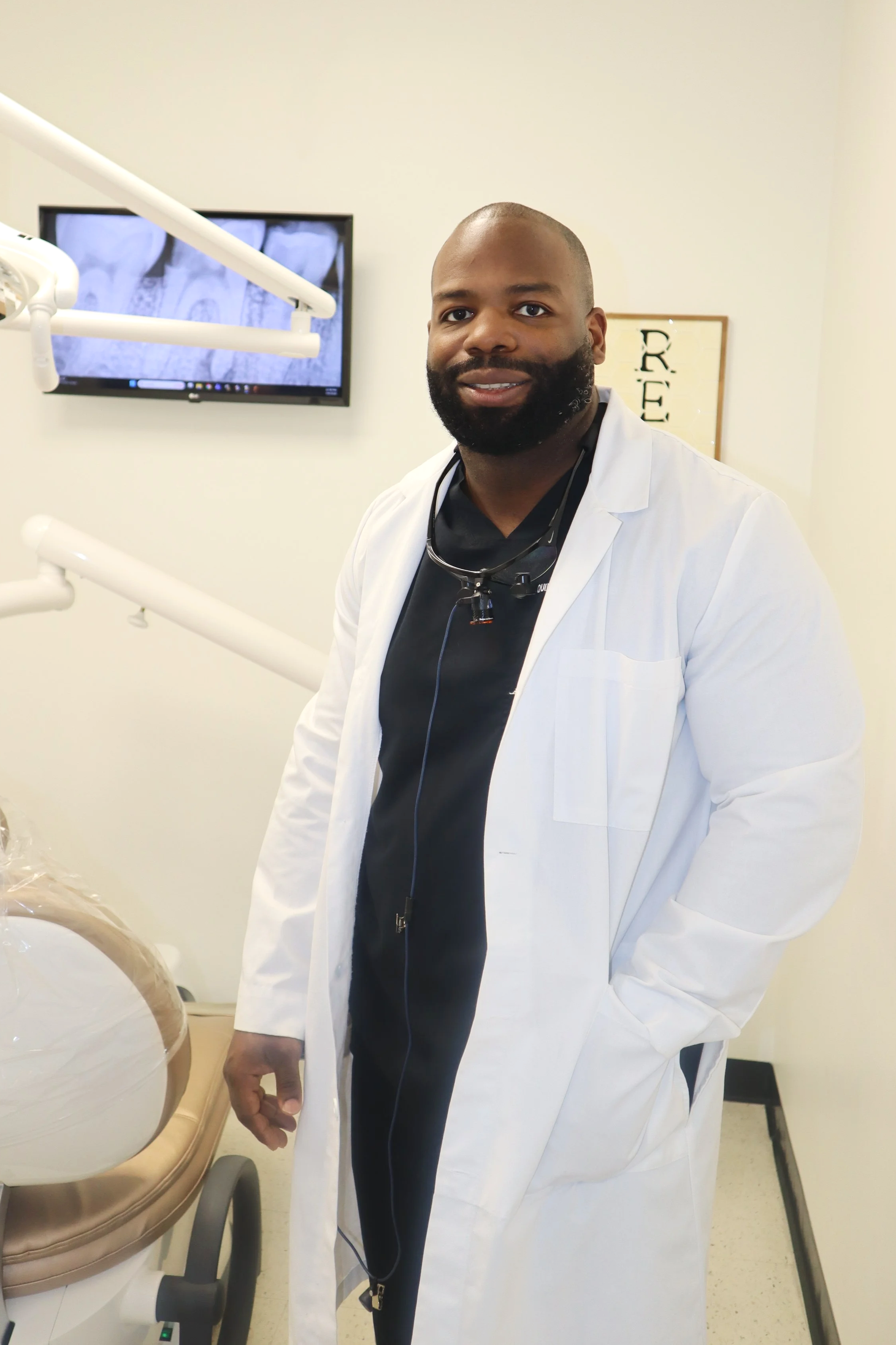 A male dentist or doctor standing inside a dental clinic, wearing a white coat, black scrubs, and a stethoscope around his neck, with dental equipment and a monitor in the background.