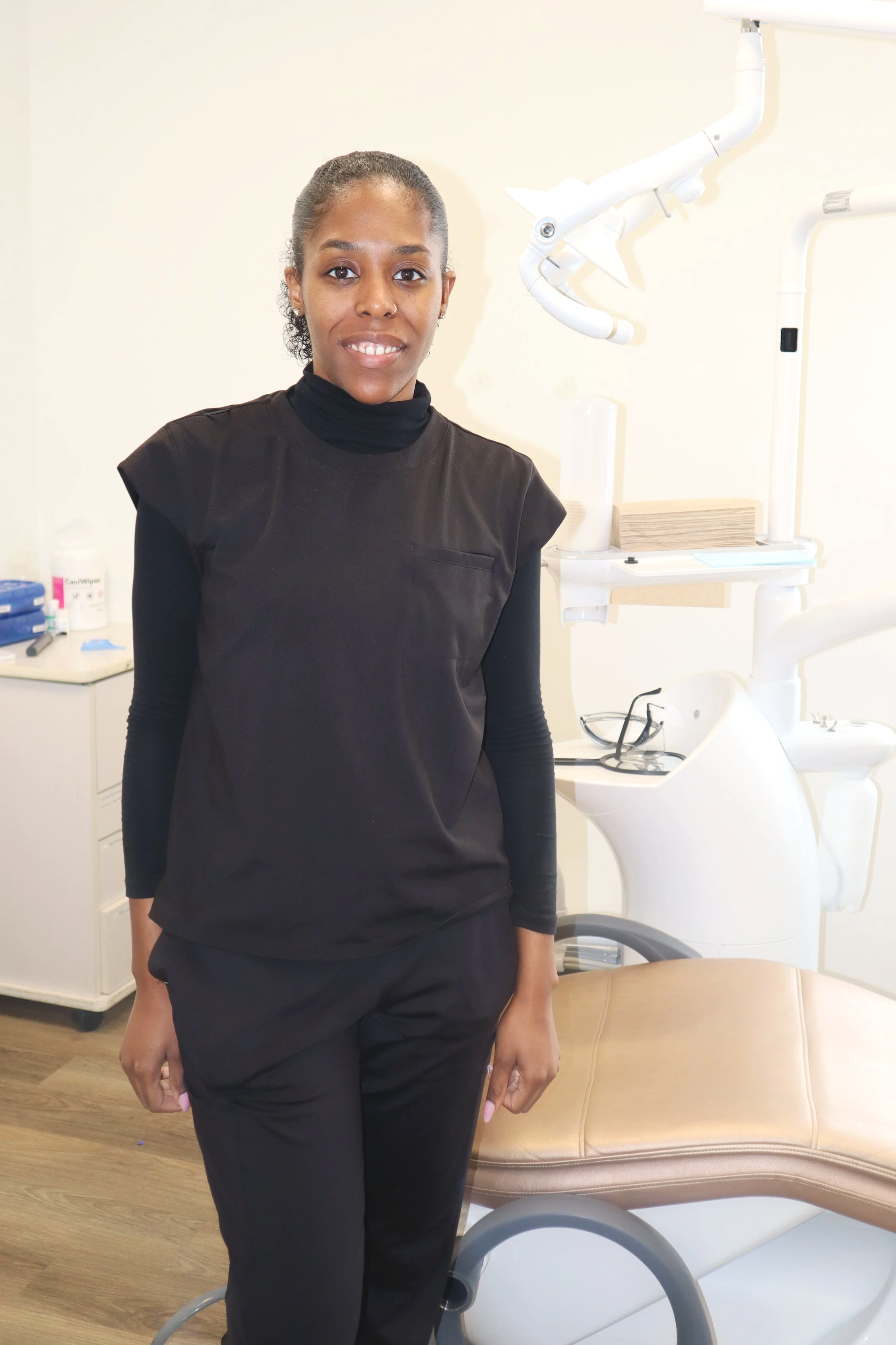 A young woman wearing black medical scrubs standing in a dental clinic with dental equipment in the background.
