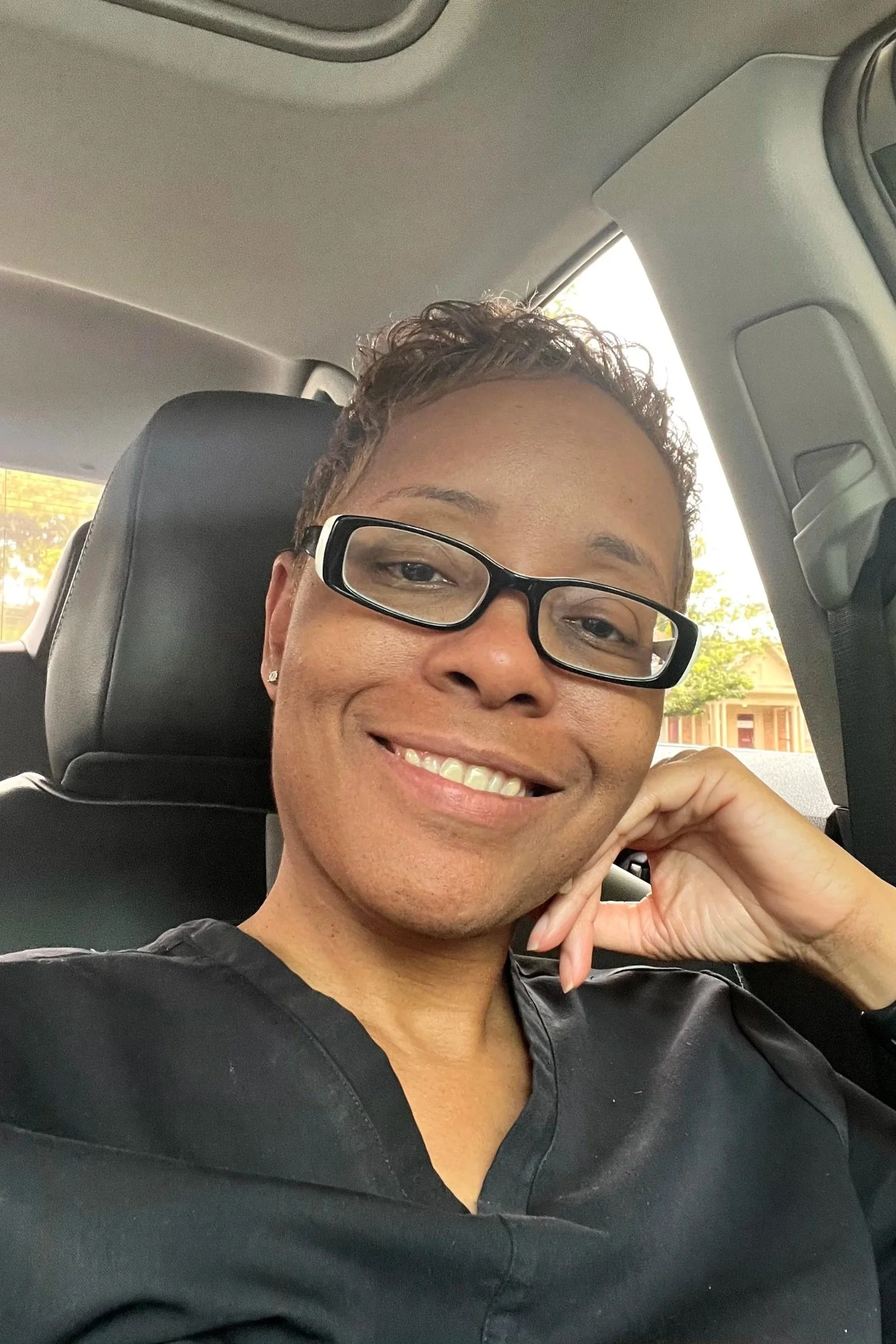 A smiling person with short curly hair, glasses, and a black shirt sitting in a vehicle.